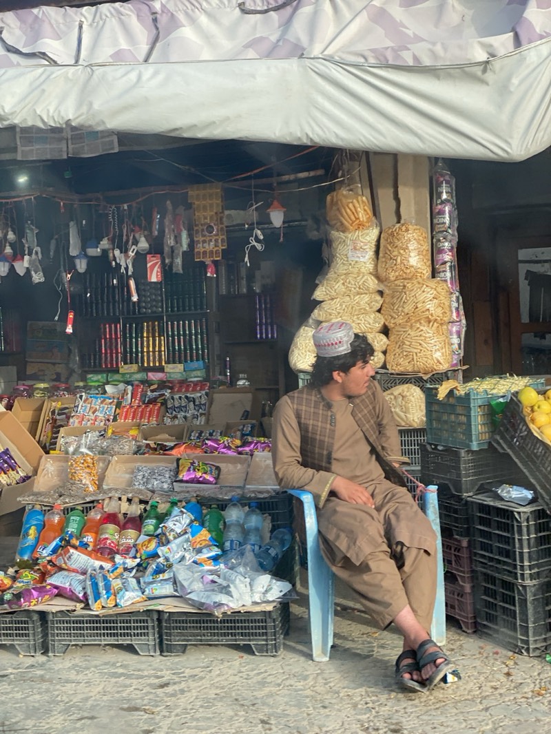 A man wearing a brown outfit and a traditional hat is seated on a blue plastic chair in front of a small shop in what appears to be Mir Ahmad Kalay, Afghanistan. The shop is well-stocked with various items including snacks, drinks, household goods, and packaged food. The shop's goods are displayed on black plastic crates. The man is looking to his right. Overhead, the shop has a striped awning and various items hanging for sale. Bags of snacks are stacked high next to the shop. Fruit is displayed in crates near the man.