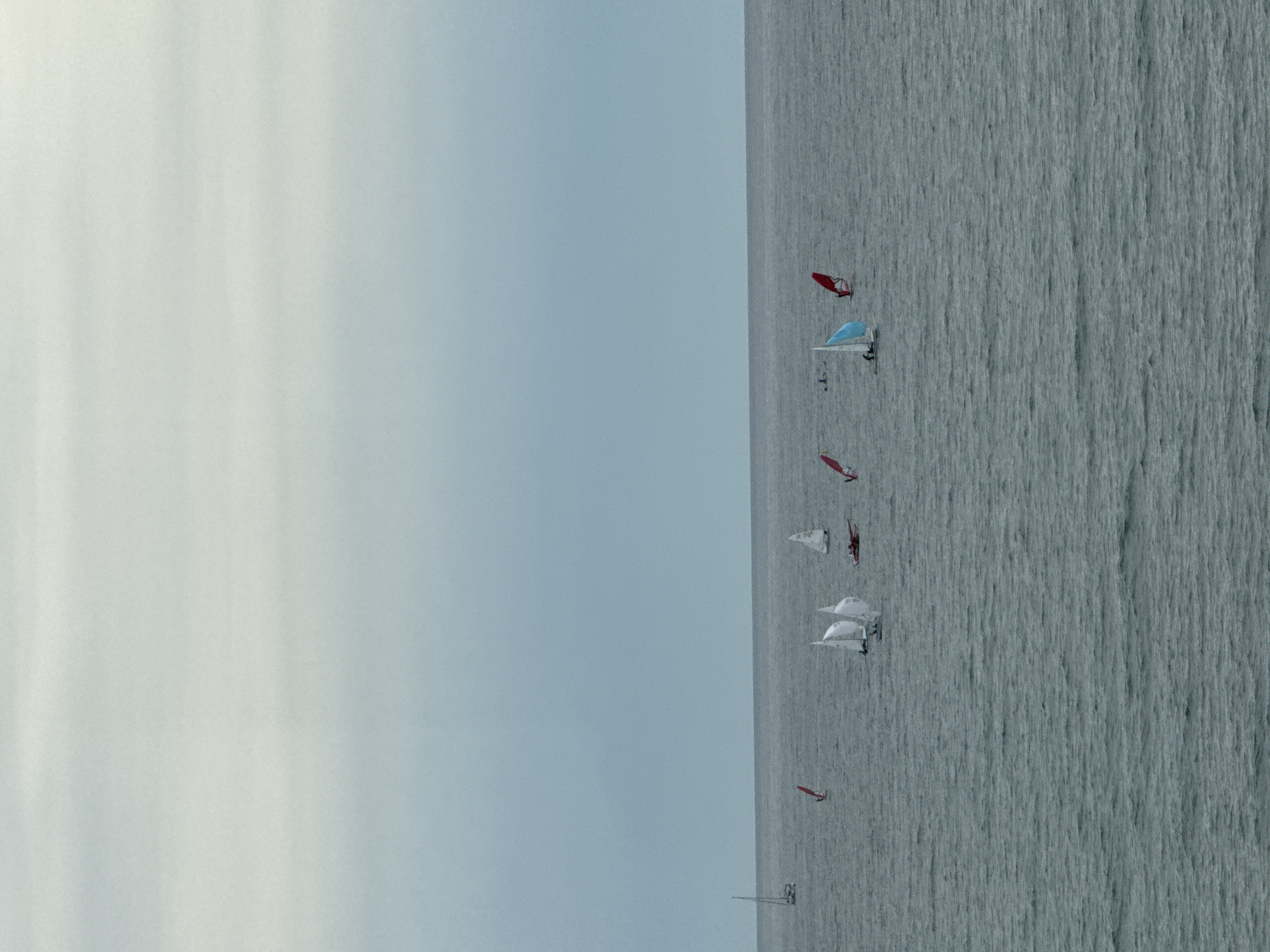 The image shows a view of the sea near Sochi, Russia, with several sailboats scattered across the water. The sky is overcast and pale. The sea appears gray and choppy. The sailboats feature sails of various colors, including white, red, and blue. One notable structure appears to be a pier or platform extending into the water.