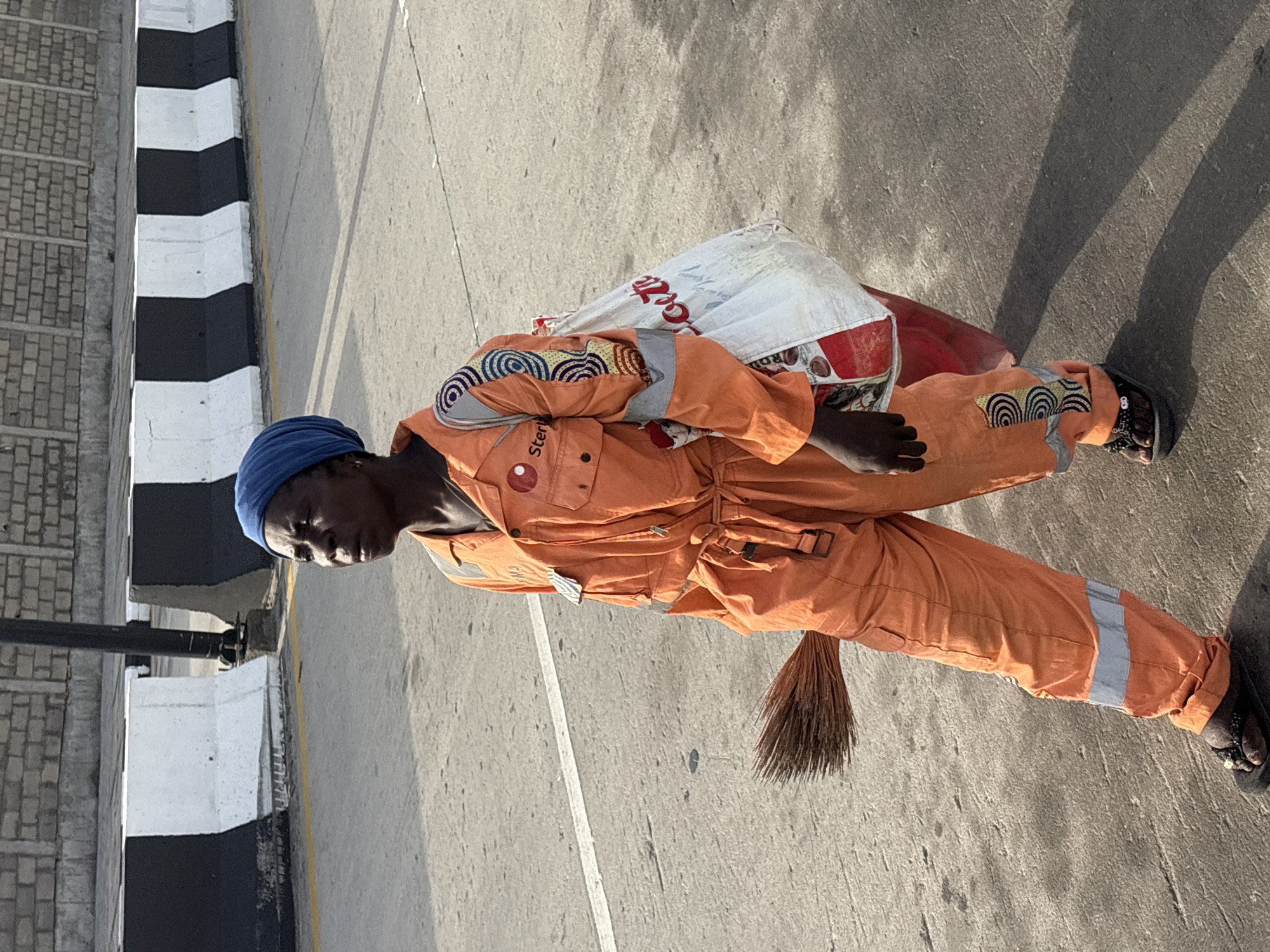 A road worker wearing an orange jumpsuit with a blue head covering stands on a concrete road, likely in Igboefon, Nigeria, as she carries a broom and a bag with the Coca-Cola logo on it. The road worker stands beside the curb, which is painted with black and white stripes.