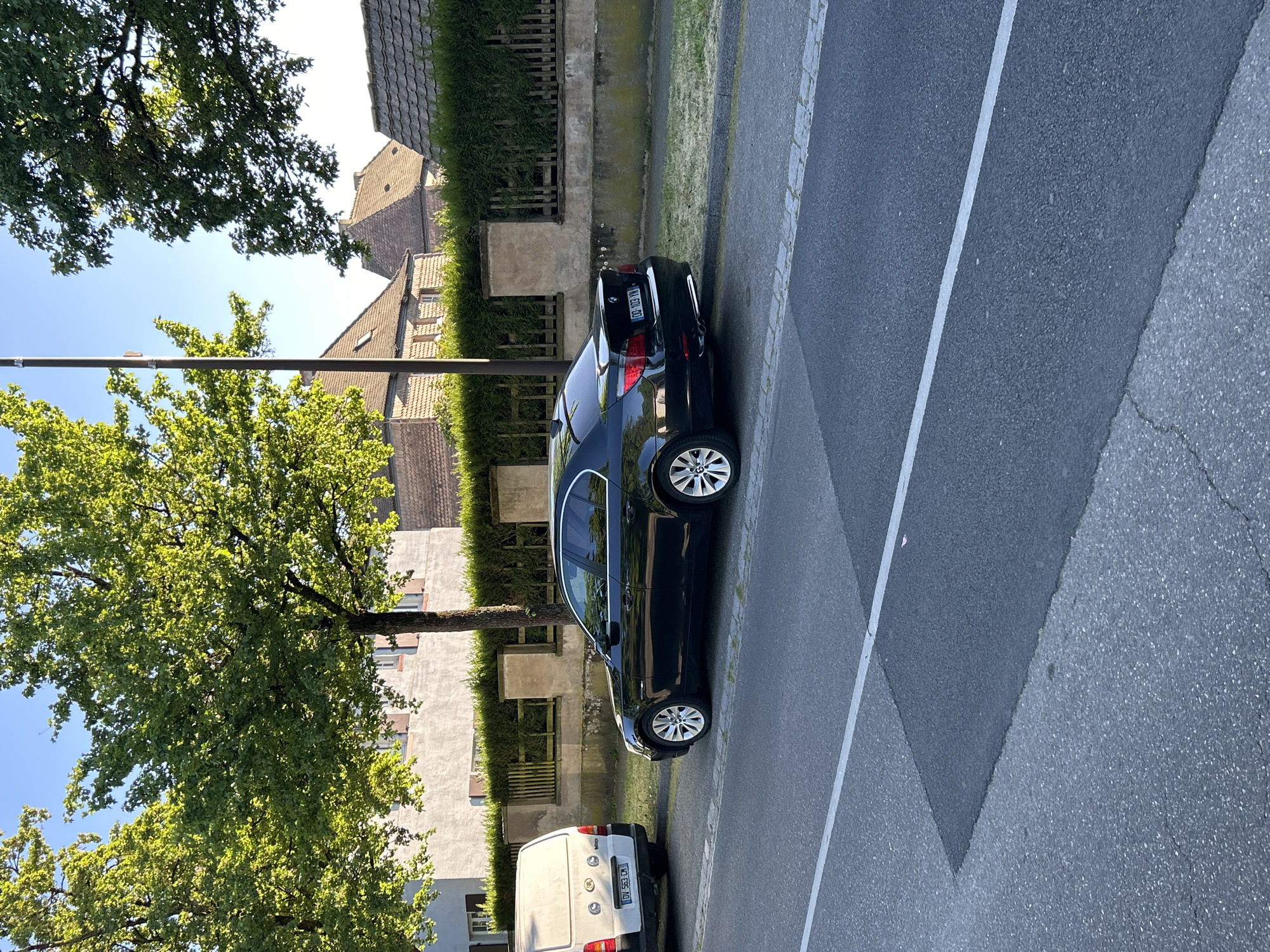 The image, taken in Strasbourg, France, depicts a quiet residential street scene under bright, clear daylight. The main subjects are two cars parked along the side of an asphalt road, flanked by buildings and lush green trees.
In the foreground, a dark-colored sedan, identifiable as a BMW by its emblem on the trunk, is parked parallel to the curb. Its rear license plate, partially visible, reads "DV-103-VN," featuring the standard French "F" on a blue strip and what appears to be the Grand Est region logo. Further down the street, behind the sedan, a white utility van is also parked. Its rear license plate displays "DV-363-CD," also with the French "F" and a regional identifier. A Renault logo is visible on the van's rear door.
To the left of the parked cars, a narrow paved area separates the road from a row of well-maintained green hedges, behind which are residential buildings. These buildings feature light-colored facades and traditional tiled roofs, some with dormer windows. Large, deciduous trees with vibrant green foliage are scattered along the street, casting shadows that suggest either morning or late afternoon sun. A blue sky with no clouds is visible through the tree canopy. The road itself is made of asphalt, with a clear white line marking a lane or parking area, and a section of cobblestone-like paving at its edge. No people or activities other than parked cars are visible.