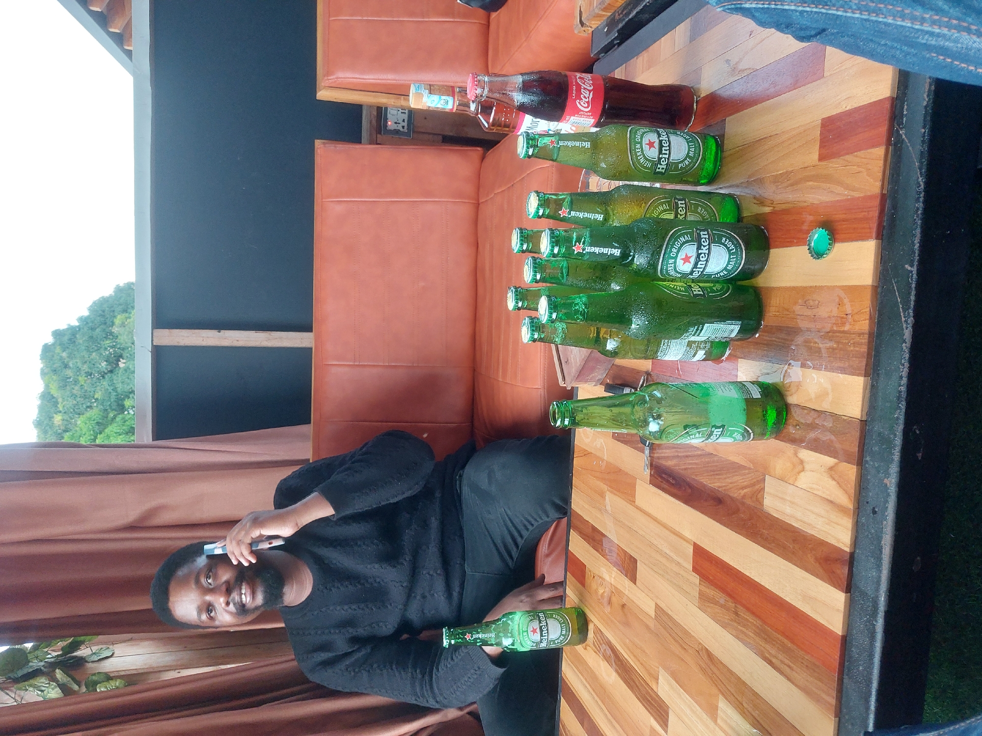 The image captures a scene at an outdoor seating area, potentially in Dar es-Salaam, Tanzania, as suggested by the trees in the background. A man is seated on a cushioned bench at a table. He is Black and appears to be talking on a mobile phone while holding a bottle of Heineken beer. The table is made of wood, and has several bottles of Heineken, a Coca-Cola bottle, and a bottle cap on it. The environment features drapery, a cushioned bench, and visible greenery outside.