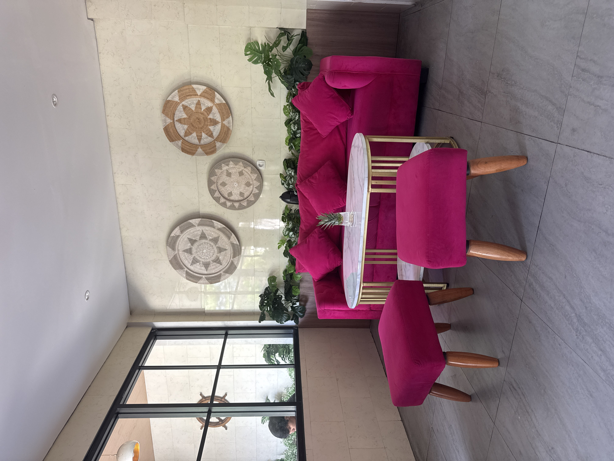 The image depicts a seating area in a brightly lit interior, possibly a lobby or lounge, located in Kuta, Indonesia.
The focal point is a bright pink velvet couch adorned with matching throw pillows. A small, oval-shaped table with a marble top and gold frame sits in front of the couch, holding a pineapple. Two matching pink velvet stools with wooden legs are positioned to complete the seating arrangement.
The wall behind the couch is decorated with three round woven wall hangings of varying sizes. Potted Monstera plants are placed behind the couch, adding a touch of greenery.
A large black-framed window allows natural light to flood the space. A decorative wooden ship's wheel is visible outside the window, along with a man looking inside.
The floor is covered in gray rectangular tiles. The overall ambiance suggests a modern, tropical-inspired design.