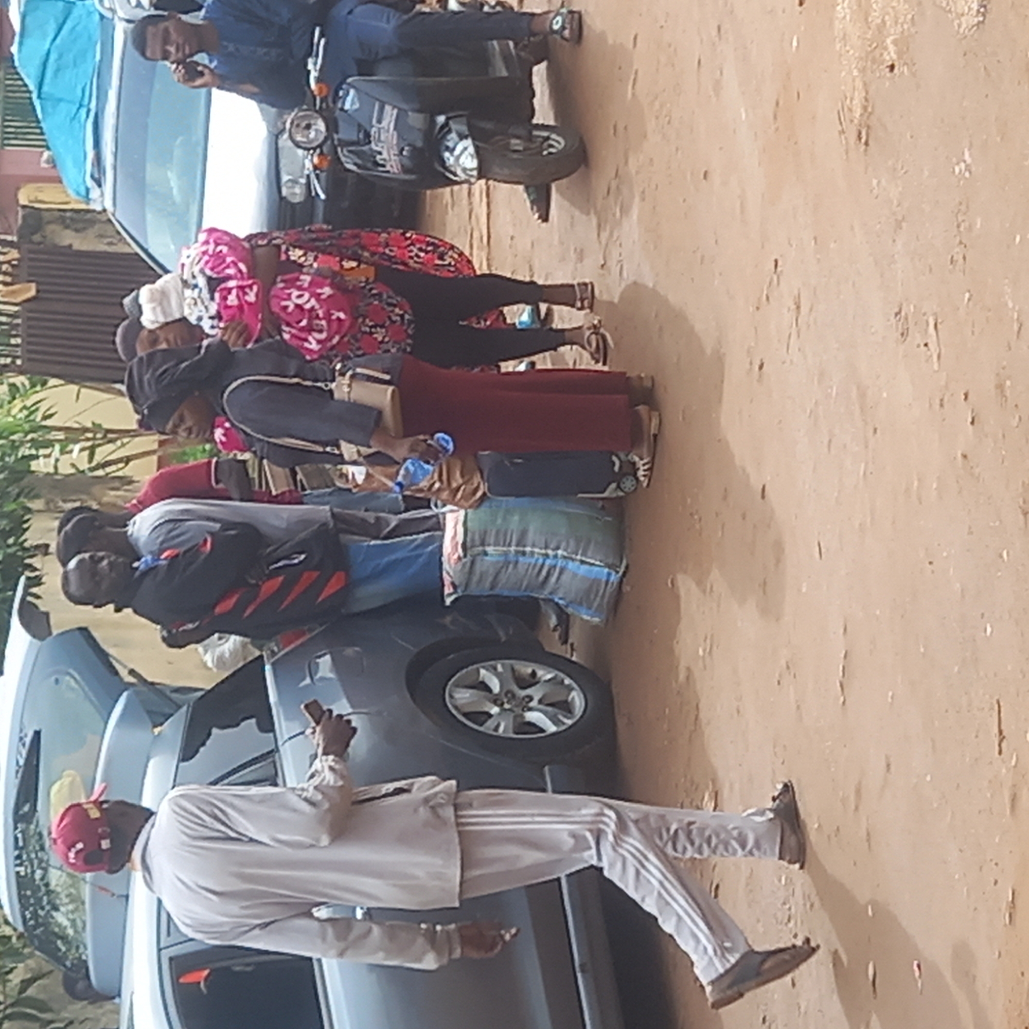 The image depicts a scene in Sardauna, Nigeria, showing a group of people gathered beside two cars and a motor scooter. A man in a white track suit and red cap stands in the foreground near one of the cars. Behind him, several people stand in a line, near bags and luggage placed on the cars. One man is on a motor scooter, using a cell phone. The environment appears to be a dusty, possibly residential street.