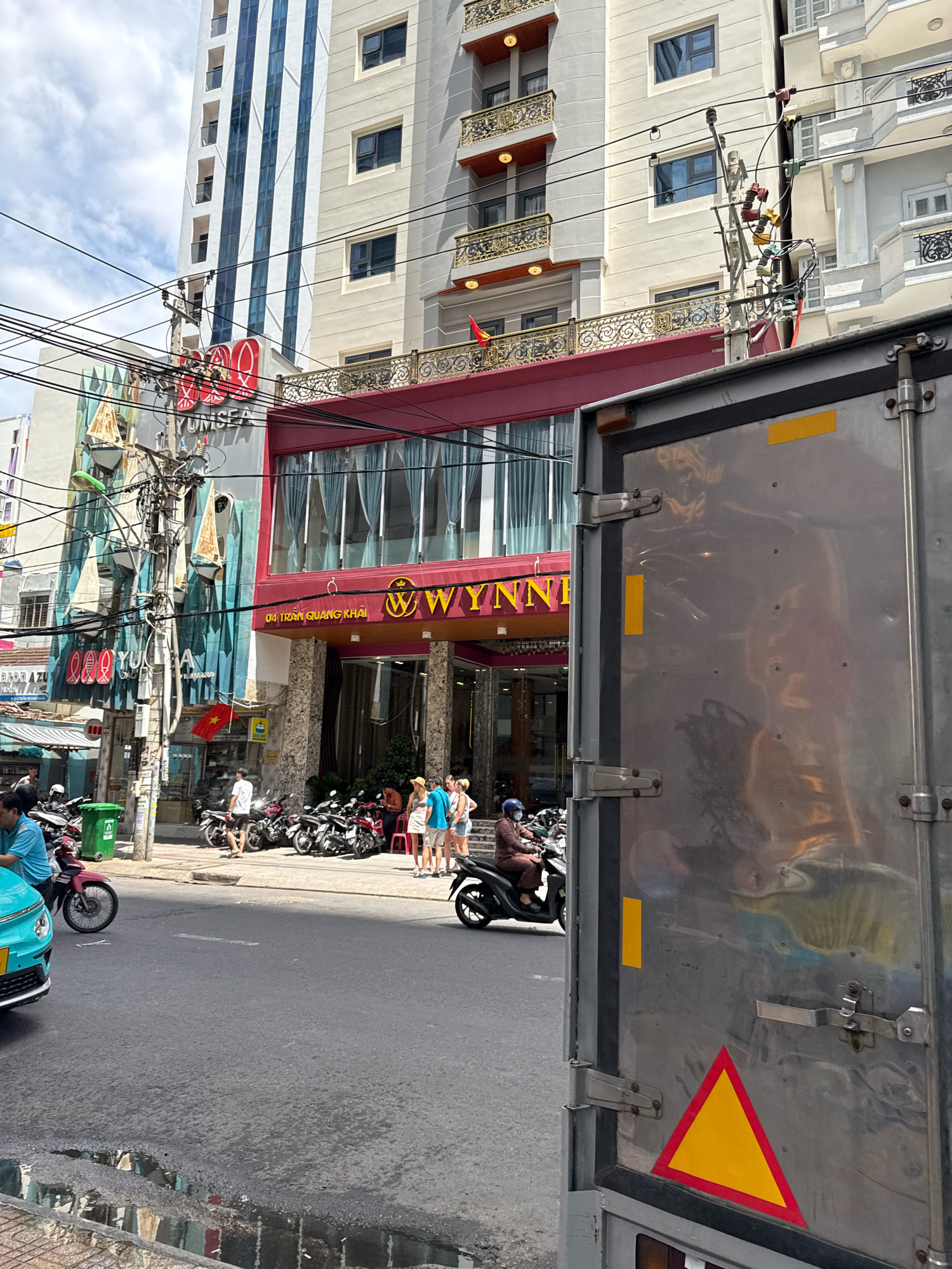 The image depicts a street scene in Nha Trang, Vietnam, with a focus on the facade of a building and a large metal truck in the foreground. The building features multiple stories with varying architectural styles. The upper floors of the central building are a light cream color, with rows of dark-paned windows and decorative balconies with gold railings. Below this, a section with dark blue vertical stripes is visible, followed by a bright red awning that stretches across the lower facade. The ground level of the building has a stone-like facade with a recessed entrance and large windows. A prominent yellow and red sign with the word "WYNN" in a stylized font is mounted above the entrance. To the left of this building, another structure is visible with a teal and white facade decorated with images of sailboats. A complex arrangement of electrical wires and poles is situated in front of these buildings.
In the street, several motorbikes are parked and in motion. A few people are visible walking and riding motorbikes. On the sidewalk in front of the Wynn building, a group of individuals is standing, some looking towards the building, others engaged in conversation. A turquoise car is parked on the left side of the street, partially obscuring a red motorbike. The road surface is asphalt, and there is a large puddle reflecting the surrounding buildings.
The foreground is dominated by the side of a large, silver metal truck. The truck's surface shows reflections of the scene and has various latches and hinges. A yellow rectangular sticker and a yellow triangle with a red border are affixed to the truck's door. The overall impression is a bustling urban environment with a mix of modern and older architectural elements, typical of a Vietnamese city.