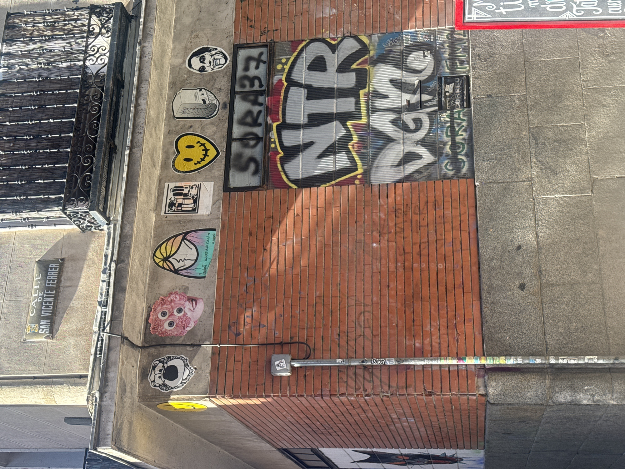 This image, captured under bright daylight, depicts an urban building facade rich with street art and architectural details, rotated 90 degrees clockwise. The scene features a multi-textured wall with sections of red brickwork, grey concrete, and glass block windows.
On the upper left, a black metal balcony with intricate, ornate railings is visible, partially obscured by a dark, textured shade. Below the balcony, a rectangular street sign reads "CALLE DE SAN VICENTE FERRER," indicating a location in a Spanish-speaking country.
The concrete sections of the wall are covered in a variety of stickers. Visible designs include a white sticker with a skull-like face, a grey sticker resembling a classical bust, and a bright yellow heart-shaped smiley face. Further down, there's a distinct pink head with multiple eyes and a colorful sticker bearing the words "NATURALEZA" and "MONOKA."
A prominent area of the wall features glass block windows, heavily covered in graffiti. Large, bold letters spell "NTR" across the center, with "SORAB7" visible to the left and "SORA FM2" partially seen elsewhere on the glass. Faint, older graffiti is also visible on the brick sections.
A metal conduit or pipe extends horizontally across the brick wall, adorned with numerous small stickers, some of which display text like "FARLOPA" and "SERO." The right side of the image shows a grey, tiled sidewalk or pavement. The clear sunlight casts distinct shadows, suggesting a sunny day in an active urban environment.