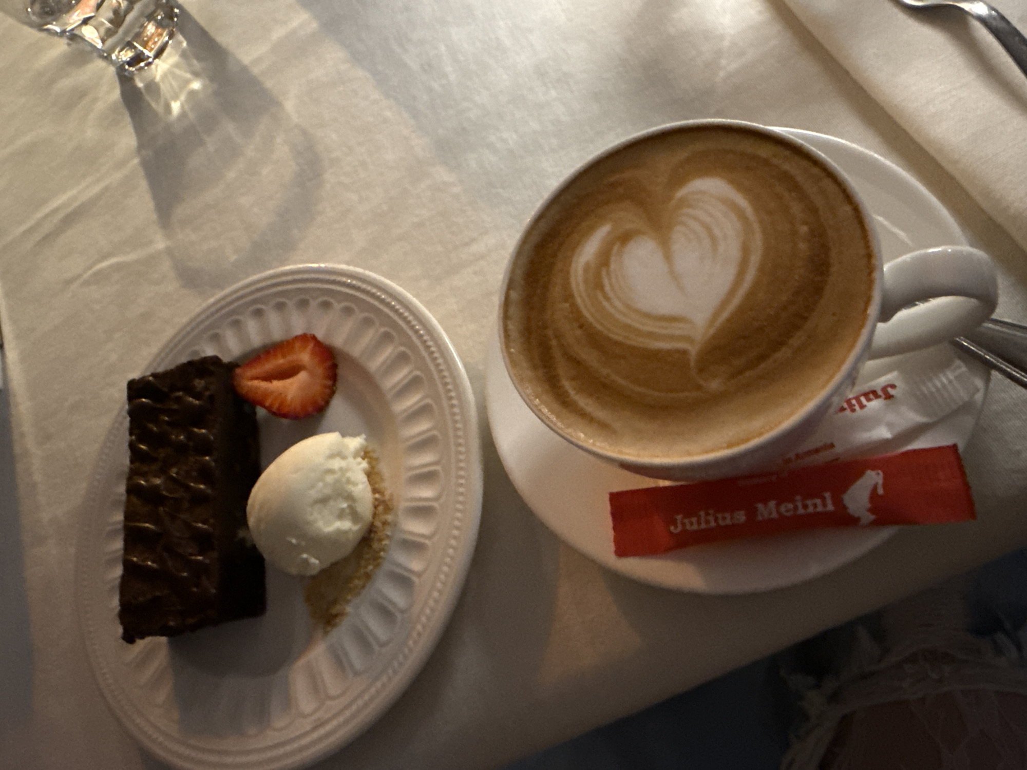 The image captures a dessert plate and a cup of coffee, possibly in a cafe in Unknown, Unknown. A rectangular chocolate brownie is accompanied by a scoop of vanilla ice cream and a sliced strawberry, arranged on a white, patterned plate. Beside the plate, a cup of coffee with latte art in the shape of a heart rests on a saucer, along with a Julius Meinl sugar packet. The scene is set on a white tablecloth, with a water glass and cutlery partially visible in the background. The lighting appears to be artificial, creating shadows and highlights on the objects.