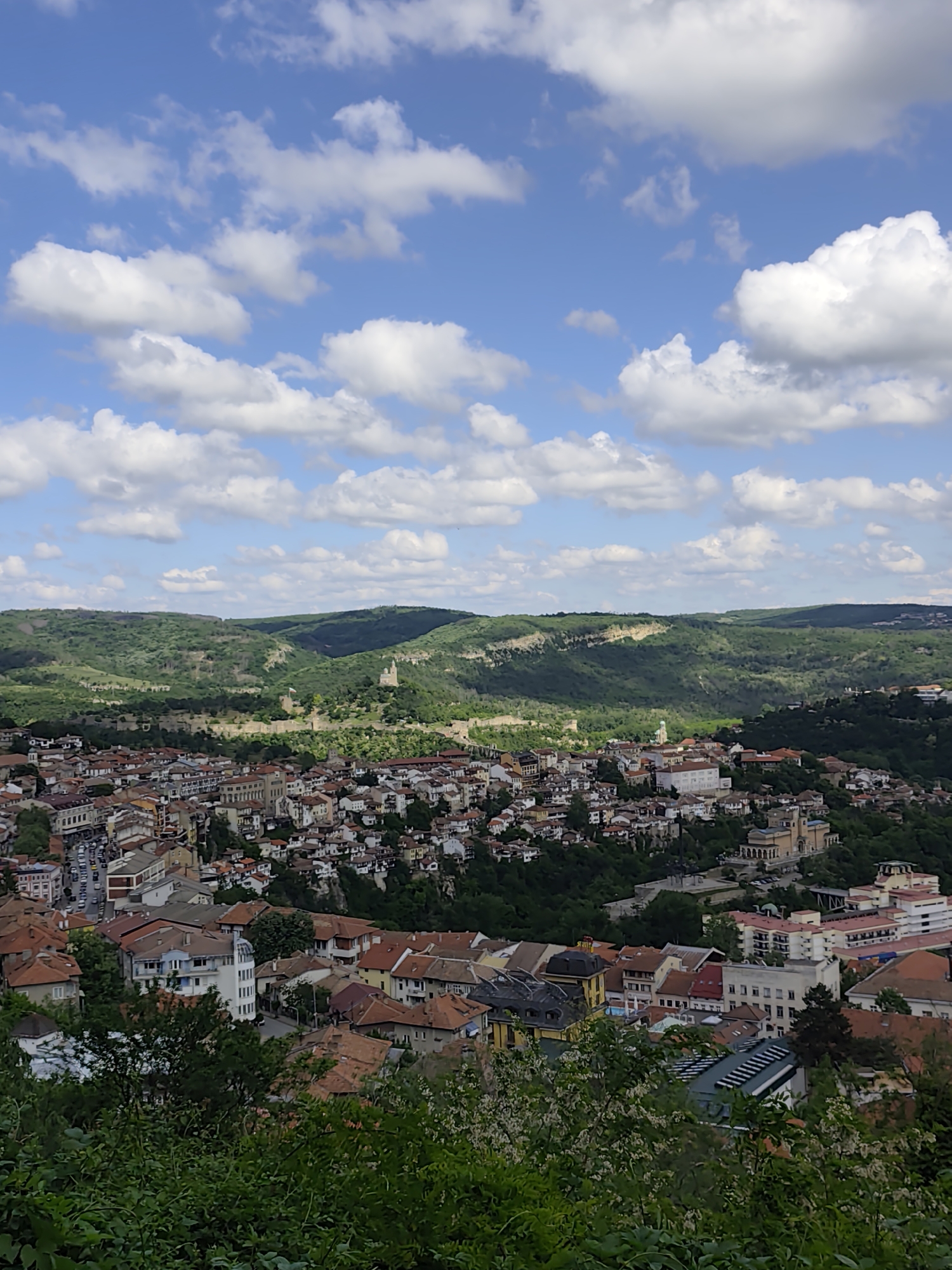 The image captures a high-angle view of a dense cityscape situated in a valley, likely in an Eastern European or Balkan region. The city consists of a multitude of buildings with red-tiled roofs, varying in height and style, interspersed with trees and greenery. In the background, a line of rolling hills covered in lush forest stretches across the horizon under a partly cloudy sky. The sky is predominantly blue with numerous white, puffy cumulus clouds spread throughout. In the mid-ground, remnants of an ancient fortification can be seen on one of the hills, hinting at a historical significance of the area. The foreground features green foliage and bushes, suggesting that the photograph was taken from an elevated vantage point, such as a hill or a high building.