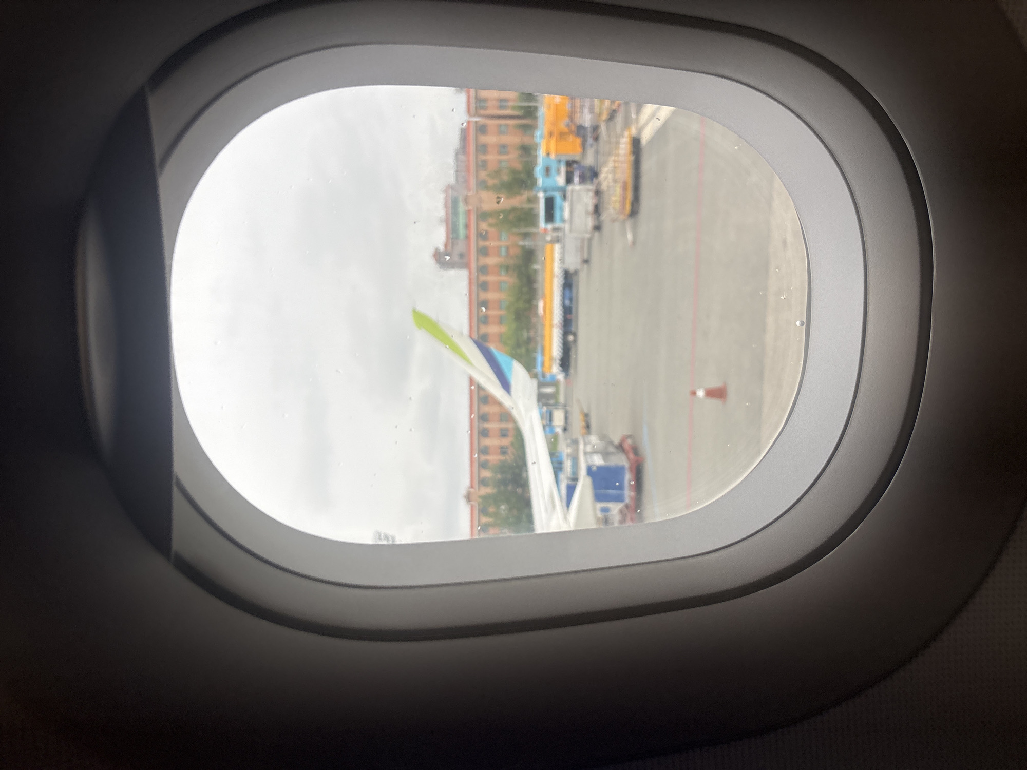 The image is taken from inside an airplane window, looking out at an airport tarmac. In the background, a large, brick building with many windows and arches is visible, likely a terminal building or hotel, situated in Busan, South Korea. The building appears to be an older architectural style. On the tarmac, an airplane wing with a green and white tip is prominent. Various airport vehicles, including trucks, baggage carts, and possibly a fuel truck, are parked or moving around. The ground is paved, and there are red and white lines indicating taxiways or parking areas. A single orange traffic cone is visible on the tarmac. The sky is overcast, and there are visible water droplets on the airplane window, suggesting it might be raining or has recently rained. The overall scene depicts a typical airport environment in an urban setting.