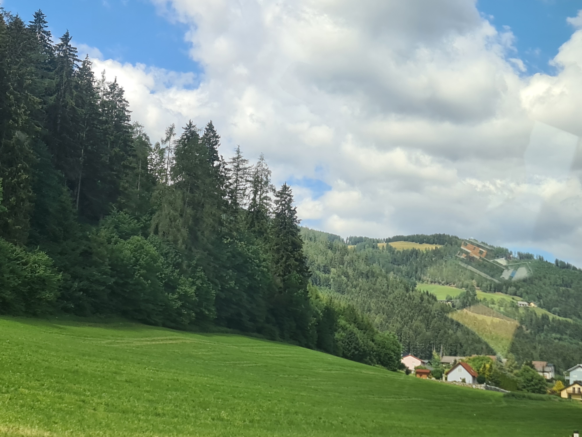 The image captures a scenic view of a green, sloping meadow in the foreground, leading up to a dense forest on a mountainside. The forest is predominantly composed of dark green coniferous trees. Beyond the forest, the landscape reveals more rolling hills covered in trees, with some patches of lighter green and a few scattered buildings. The sky above is a mix of blue patches and fluffy white clouds. In the distance, on the upper part of the mountain, some structures are visible, possibly ski jumps or other recreational facilities, indicating the location is likely near St. Peter ob Judenburg, Austria, a known area for winter sports. Several small houses with red and white roofs are nestled amongst the trees on the lower slopes. The overall impression is one of a peaceful rural mountain setting.