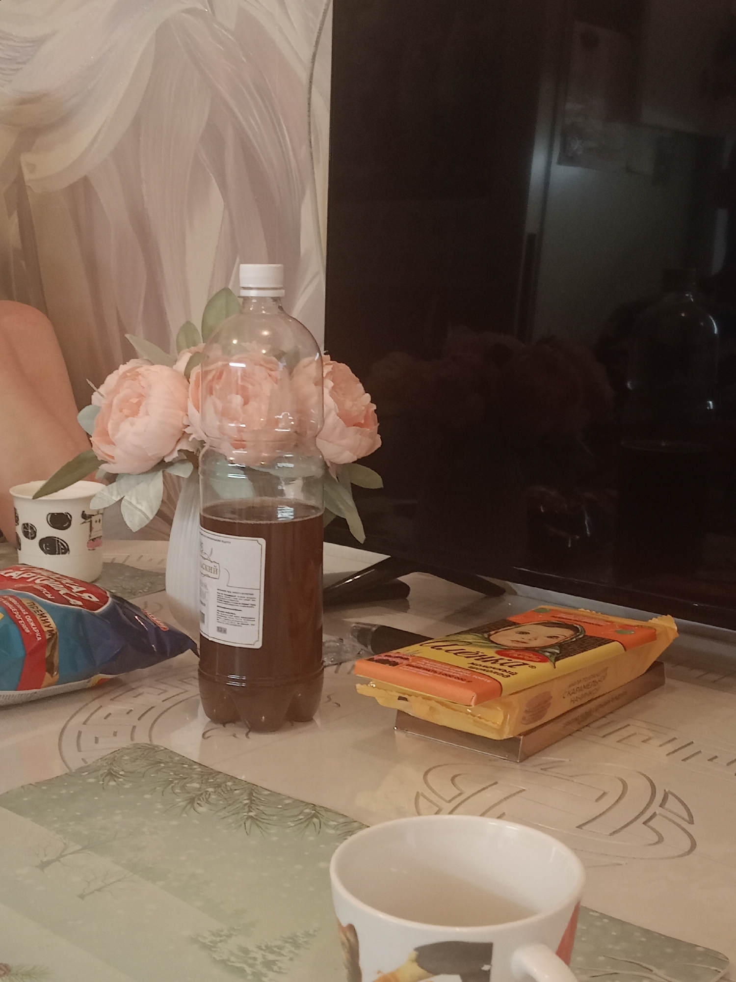 This media file captures a still life arrangement on a table, likely in a domestic setting in Roshchino, Russia. The primary subjects are objects arranged on a tablecloth with a light, decorative pattern.
In the center of the frame, a clear plastic bottle filled with a dark liquid, possibly a beverage, stands prominently. A white label with text is affixed to the bottle. To the left of the bottle, a bouquet of soft pink flowers, possibly peonies, is displayed in a white vase. Beside the flowers, a white paper cup with a black dotted pattern is visible. In the foreground, a partially unwrapped bag of snacks, with red and blue packaging bearing some text, lies on the table. Further to the right, a yellow package with an image of a child's face and Cyrillic text is stacked on another thin package. A white ceramic mug with a handle and a partial image on its side is in the lower right corner. In the background, a large, dark television screen is positioned behind the objects. The overall lighting suggests an indoor setting, possibly in the evening or with artificial light, and the atmosphere feels casual and domestic. The presence of a TV and the arrangement of snacks and drinks hint at a relaxed or leisure activity.