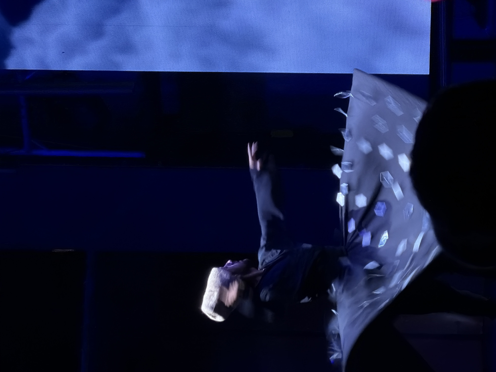 A performer, possibly a Sufi dervish, is captured in mid-spin during a dance performance. They are wearing a tall, tan, cylindrical hat and a dark robe that flares out, adorned with a pattern of light-colored geometric shapes. The performer's head is blurred due to the rapid motion, but their face is visible in profile, looking upwards. Their right arm is extended outwards, also showing motion blur. The setting appears to be a stage with dark blue walls and a large, illuminated blue screen on the right side, displaying a cloudy, ethereal image. The lower part of the image shows the dark silhouettes of audience members. The scene suggests a cultural or religious performance taking place in Manavgat, Turkey.