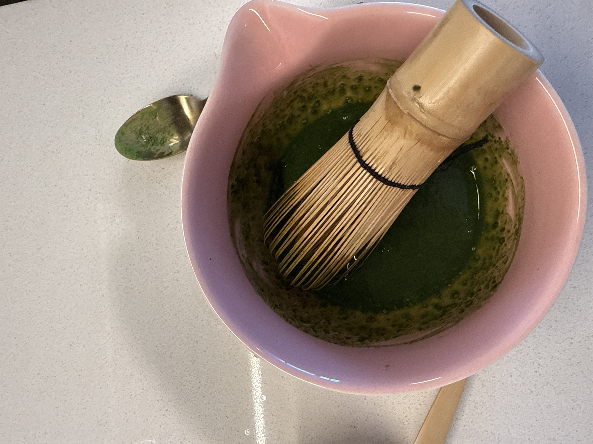 A close-up shot shows a pink ceramic bowl filled with whisked matcha tea. A bamboo matcha whisk, known as a chasen, rests in the bowl, its tines submerged in the vibrant green liquid. The matcha has a frothy, textured surface with small bubbles. To the upper right of the bowl, a small golden spoon with green residue is visible. The bowl and spoon are placed on a speckled, light-colored countertop. The overall scene suggests the preparation of a traditional matcha beverage. No specific location in Dubai, United Arab Emirates can be determined from the image.
