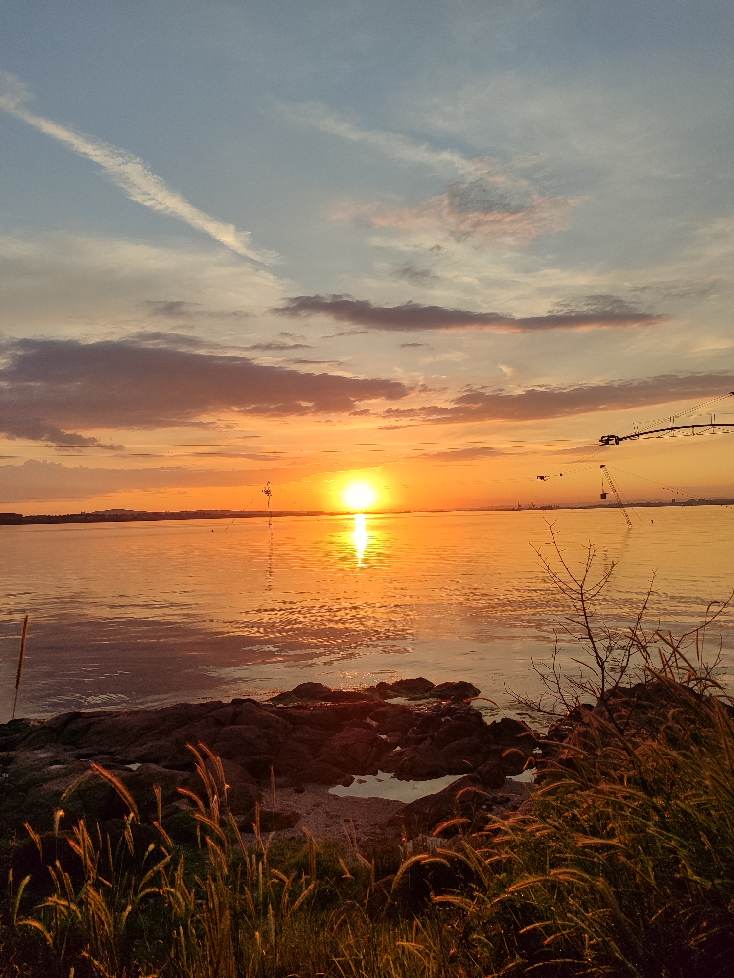 The image captures a sunset over the water near Burgas, Bulgaria. The sun is setting on the horizon, reflecting intensely on the water's surface. Clouds are present in the sky, exhibiting shades of orange, pink, and grey. In the foreground, there is vegetation, including grasses and bushes, along a rocky shoreline. Further out in the water, structures resembling cranes or pylons are partially submerged, indicating a possible industrial or harbor area. The horizon shows a faint outline of land.