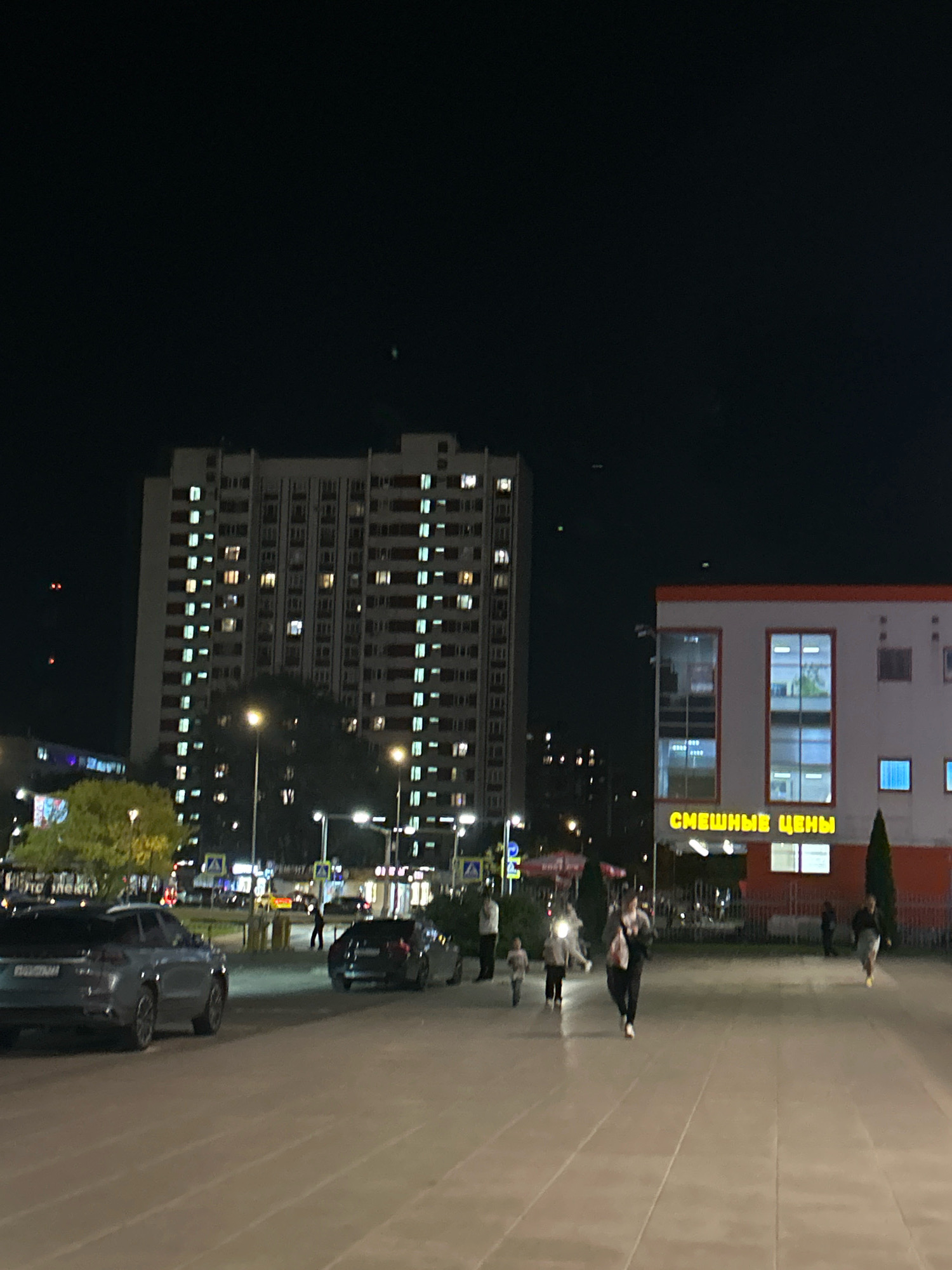 The image depicts a nighttime street scene in Moscow, Russia, with a focus on a residential building and a commercial establishment. A tall, multi-story apartment building with numerous illuminated windows dominates the left side of the frame. In front of it, trees and streetlights are visible. To the right, a low-rise building with a sign displaying yellow Cyrillic text, "СМЕШНЫЕ ЦЕНЫ" (which translates to "Funny Prices"), is illuminated. Several cars are parked on the street, including a silver SUV in the foreground. A few people are walking on the paved area in front of the commercial building, some appearing to be adults and children. The overall atmosphere is that of a typical urban environment at night.
