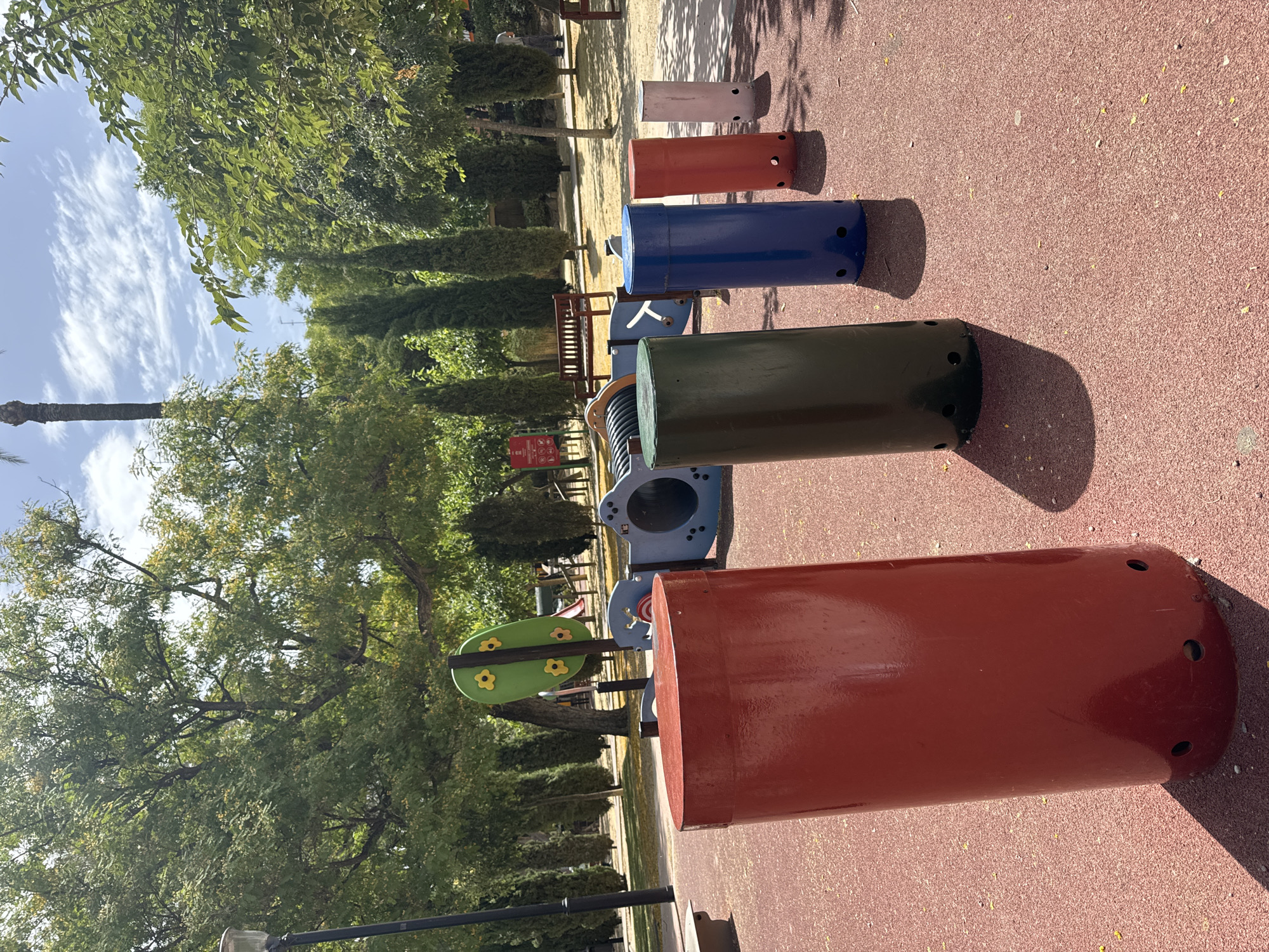 The image depicts a playground in the Jardín de Floridablanca, Murcia, Spain. In the foreground, five cylindrical structures are arranged in a line on a red rubberized surface. From left to right, these structures are red, dark green, blue, orange, and pink. The red cylinder is the largest and closest to the viewer. Behind these, partially visible, is a blue and grey play structure with tunnels. Further back, a wooden bench sits under trees. The background is filled with lush green trees, some of which are tall and slender cypress trees, interspersed with deciduous trees and a tall palm tree. The sky above is blue with scattered white clouds. A lamppost is visible on the far left. The scene is illuminated by bright sunlight, casting shadows from the cylinders and trees. No people are clearly visible in the image.