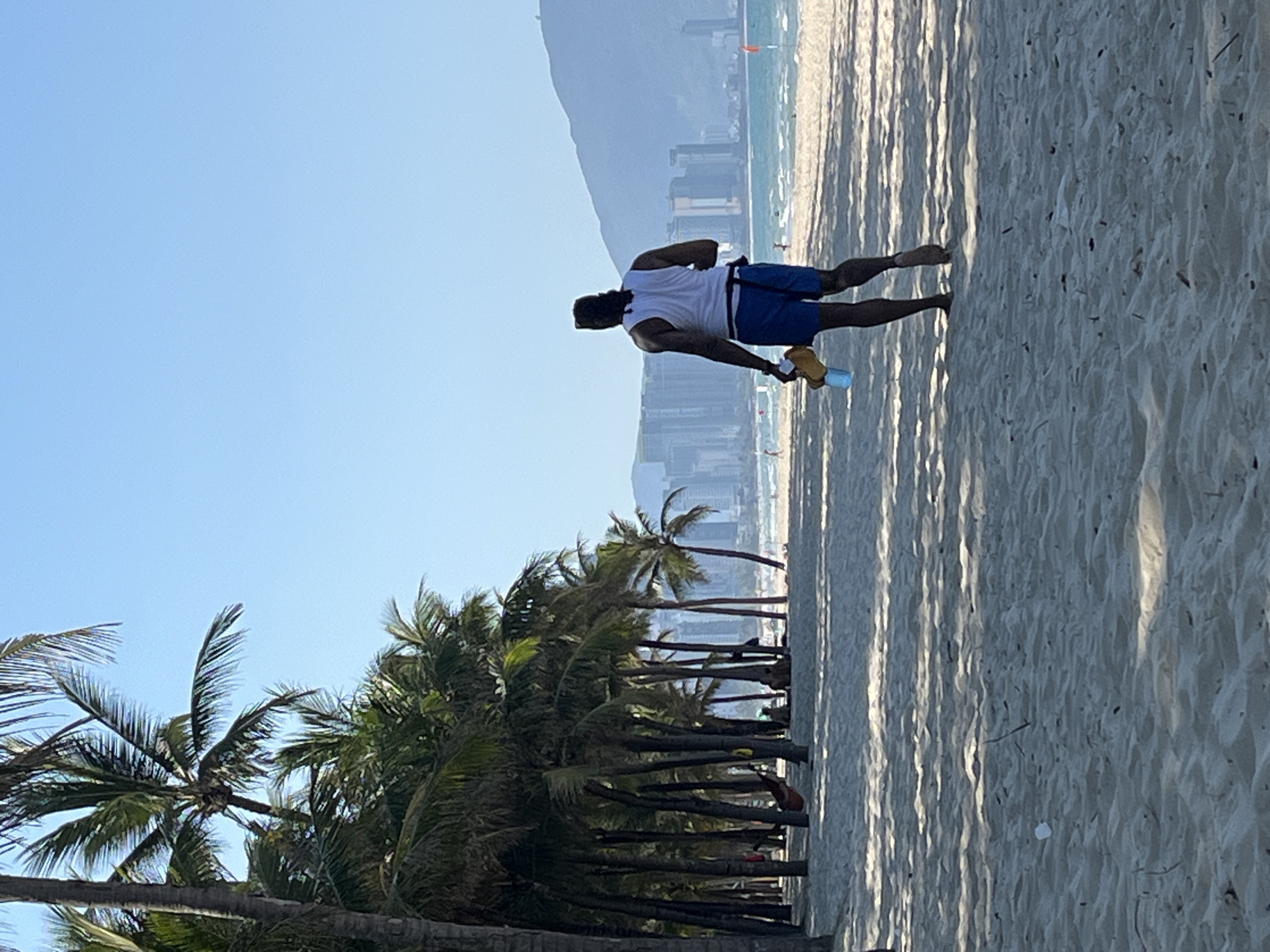 A medium shot depicts a man walking barefoot on a sunny beach with palm trees in Đà Nẵng, Vietnam. The man, facing away from the viewer, is wearing a white tank top, blue shorts, and has a dark complexion. He appears to be holding a water bottle in his left hand. The beach has a sandy texture with visible shadows cast by the palm trees. In the background, a mountain range can be seen along with the city skyline. The sky is a clear, light blue. The scene is oriented horizontally.