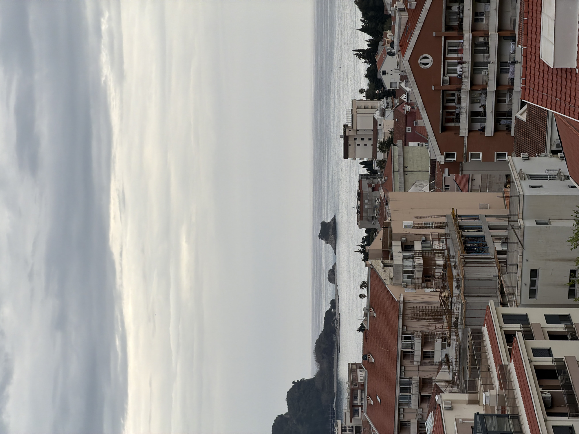The image depicts a cloudy cityscape with a body of water in the middle. The right side of the image is filled with buildings of varying heights and architectural styles, primarily featuring red-tiled roofs and apartment-style windows. Some buildings appear to be under construction. The left side shows a cloudy sky and overcast water. There are also several small islands in the water. The location is likely Budva, Montenegro, given the architecture and proximity to the Adriatic Sea.