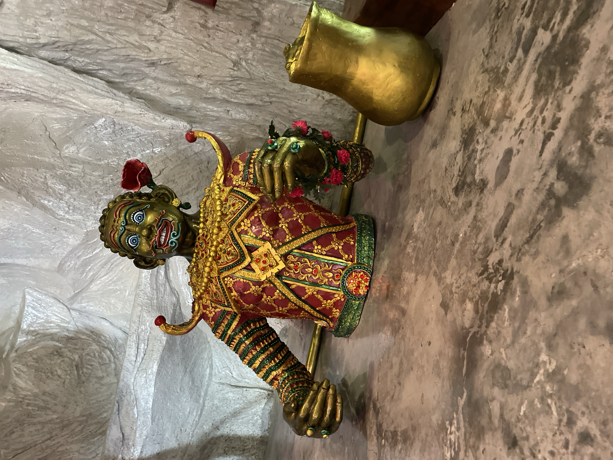 The image shows a statue of a figure, possibly a deity or mythical character, in a richly decorated costume. The figure has a green face with prominent blue and red markings around the eyes and mouth, giving it a fierce or theatrical appearance. It wears an elaborate gold and red garment adorned with intricate patterns and red jewels. A red flower is tucked behind its ear. The statue is positioned in front of a textured, light-colored wall, which appears to be made of sculpted material resembling rock or stucco. To the right of the statue, there is a golden, pitcher-shaped object. The statue is placed on a marble-like surface, which extends into the foreground. The lighting suggests an indoor setting. The location is identified as Bo Win Subdistrict, Thailand.