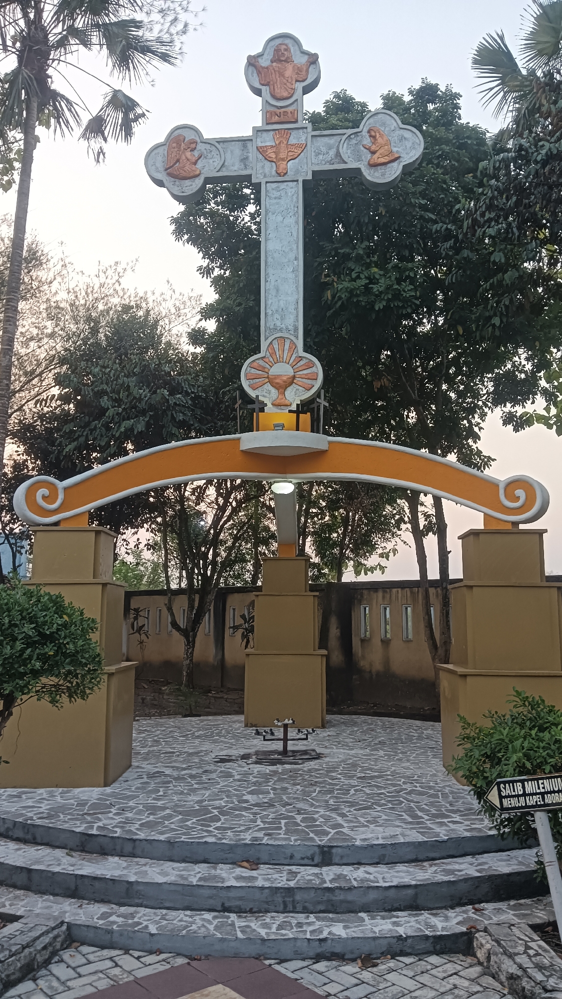 The image shows a large, ornate cross monument situated outdoors in what appears to be a public space or religious site in Dukuh, Indonesia. The cross itself is made of concrete and features decorative elements, including figures of Jesus, an angel, a dove, and possibly other religious symbols, rendered in a contrasting orange color. The cross is mounted on a stone pedestal and framed by an arched structure with curved decorative ends. Below the arch, there are several square, stepped stone structures and a small metal stand with what look like candle holders. The ground is paved with irregular stone tiles. In the background, there is a wall with small rectangular windows, trees, and palm trees. A sign pointing to the right with text "SALIB MILENIU" and "MENUJU KAPEL ADORA" is visible in the lower right corner. The lighting suggests it is either early morning or late afternoon. There are no people visible in the image.
