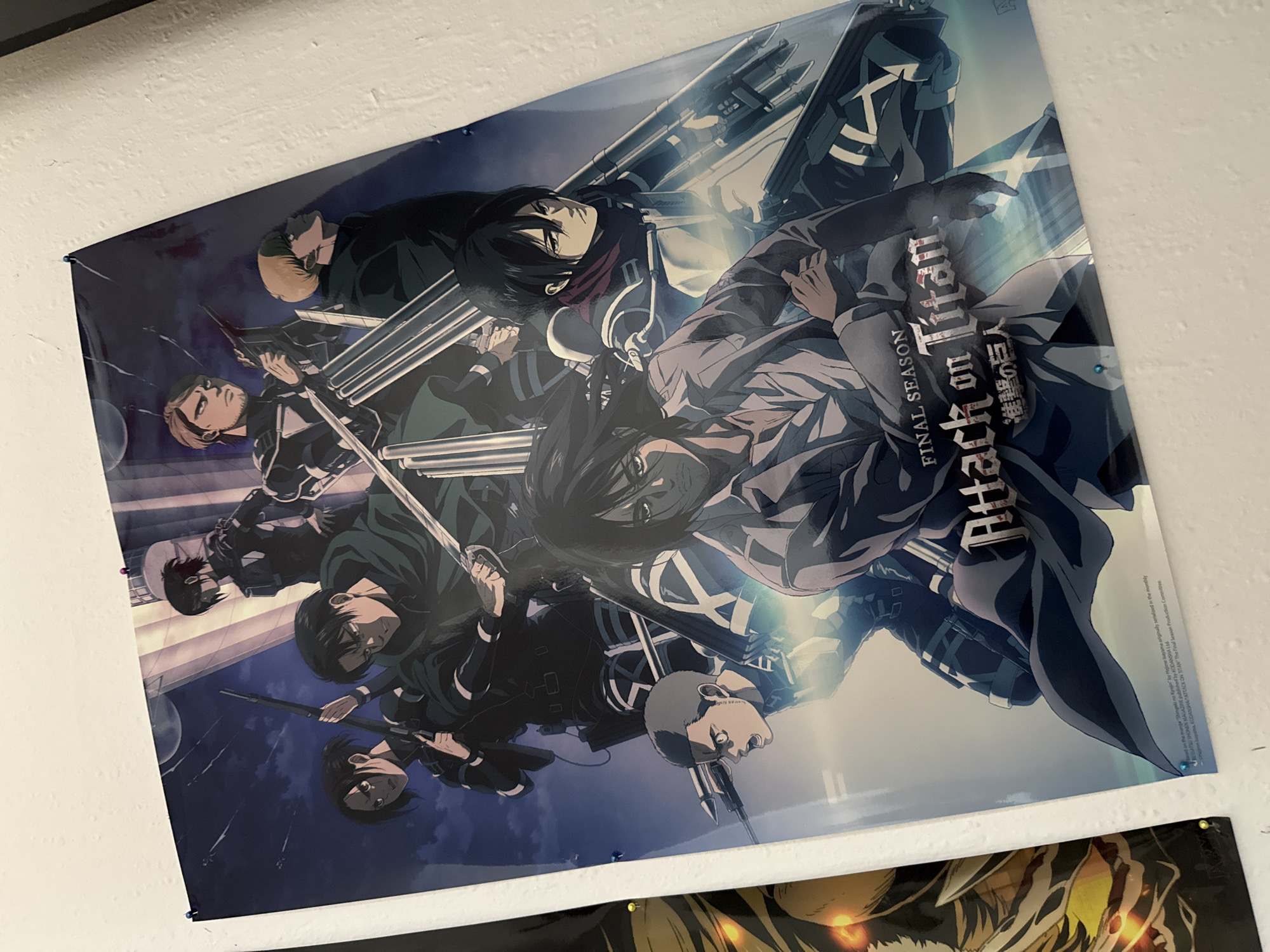 A poster is affixed to a white, textured wall with thumbtacks. The poster depicts characters from the anime "Attack on Titan." In the foreground, a central figure with dark hair and a light-colored shirt is prominent, looking forward. To the left, a character with short, dark hair and an intense expression is visible. To the right and slightly behind, a character with dark hair, wearing a red scarf, gazes forward. In the middle ground, several other characters are present, including one with a green cloak and sword, and another with short blonde hair. The background features a dark sky with streaks of light and architectural elements. The poster is glossy and reflects light in some areas. This scene is located in Bad Sachsa, Germany.