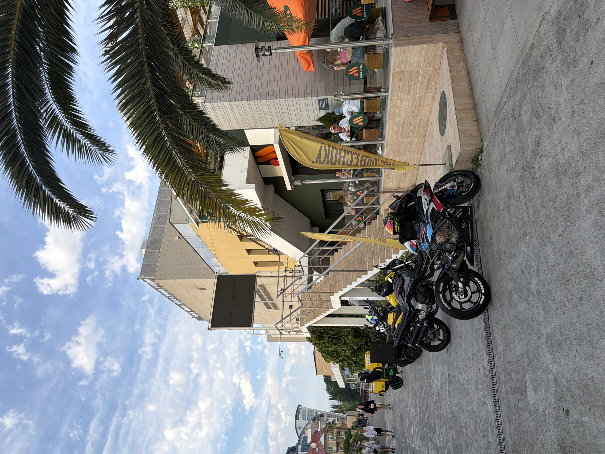 The image depicts a street scene in Sochi, Russia, likely during daylight hours, with a clear sky and scattered clouds. A multi-story building with a modern architectural style, featuring a beige facade and balconies, dominates the background. In front of the building, an outdoor seating area of a cafe or restaurant is visible, with patrons seated at tables. A prominent yellow flag with text is planted near the entrance.
In the foreground, several motorcycles are parked along the paved walkway. These include a sports motorcycle with a distinctive blue, black, and white design, and a yellow scooter. A few people are walking on the sidewalk to the left of the frame, and others are seated in the outdoor cafe. Palm trees are present in the scene, suggesting a warm climate characteristic of Sochi. The overall impression is one of a commercial area, possibly a promenade or a street adjacent to a beach resort.