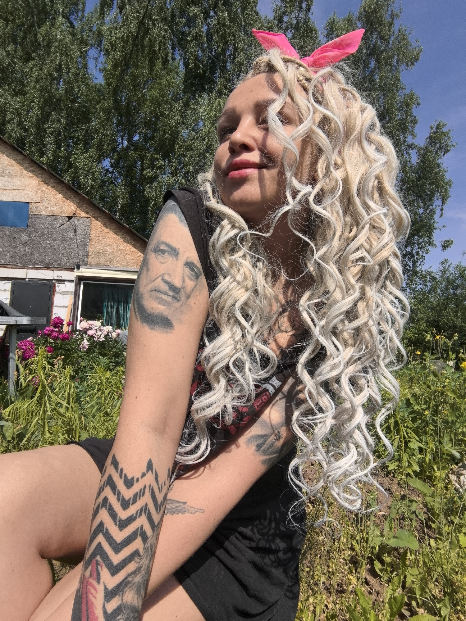 A Caucasian woman with blonde, curly hair styled in braids and a pink bow sits outdoors in a sunny, rural location. She wears a black t-shirt. Her left arm has a prominent tattoo of a man's face on the upper part, and a geometric zigzag pattern on the forearm. Her right arm shows a tattoo of a bird. The background consists of greenery, trees, and a rustic, unfinished building. The image appears to be taken in or around Noviy, Russia, as indicated by the provided information.