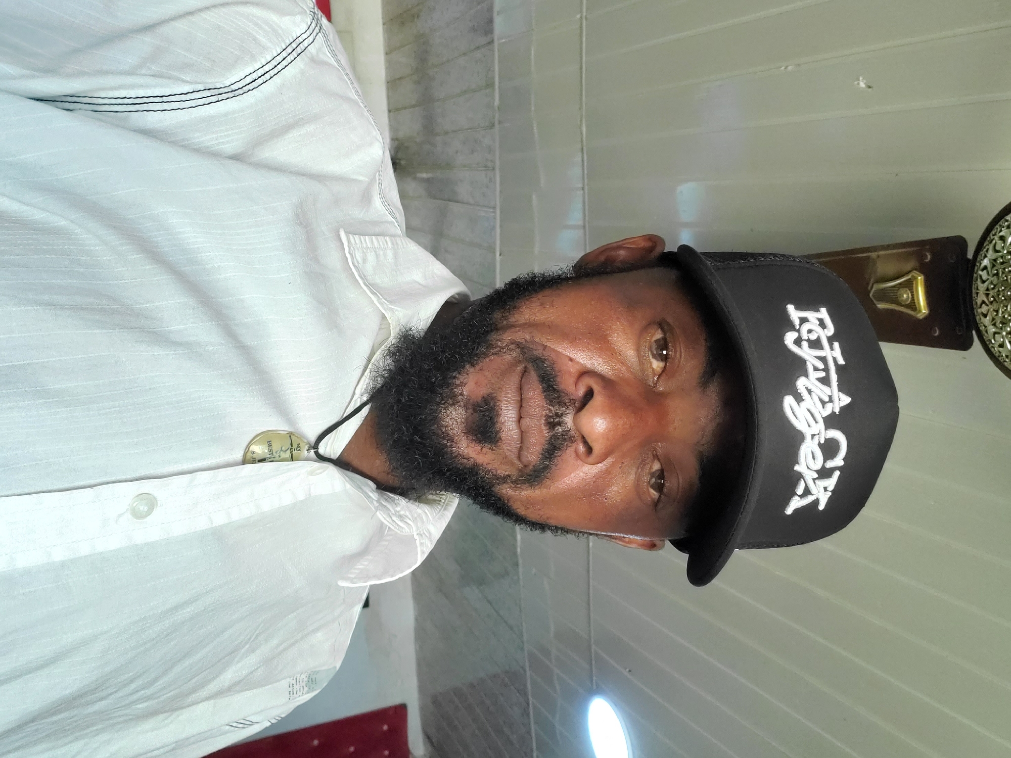 The image, rotated vertically, features an adult man with dark skin and a full black beard, looking directly at the camera. He wears a white collared shirt with subtle horizontal textured patterns and black stitching visible on the upper left side. Around his neck, a round, metallic-colored pendant hangs on a black cord, with partially visible text including "BEST" and possibly "5.15". He is also wearing a black baseball cap with white embroidered text that clearly spells "Faygo" in a stylized font, followed by what appears to be "OK". The man has a neutral to slightly pleasant expression.
The setting is indoors. Behind him, on the viewer's left (when the image is correctly oriented), is a light-colored wall with horizontal paneling or tiling. On the right, an ornate metallic object, possibly a decorative fixture or part of a mirror frame, is visible against a darker background. A bright light source is present in the lower right corner of the frame.