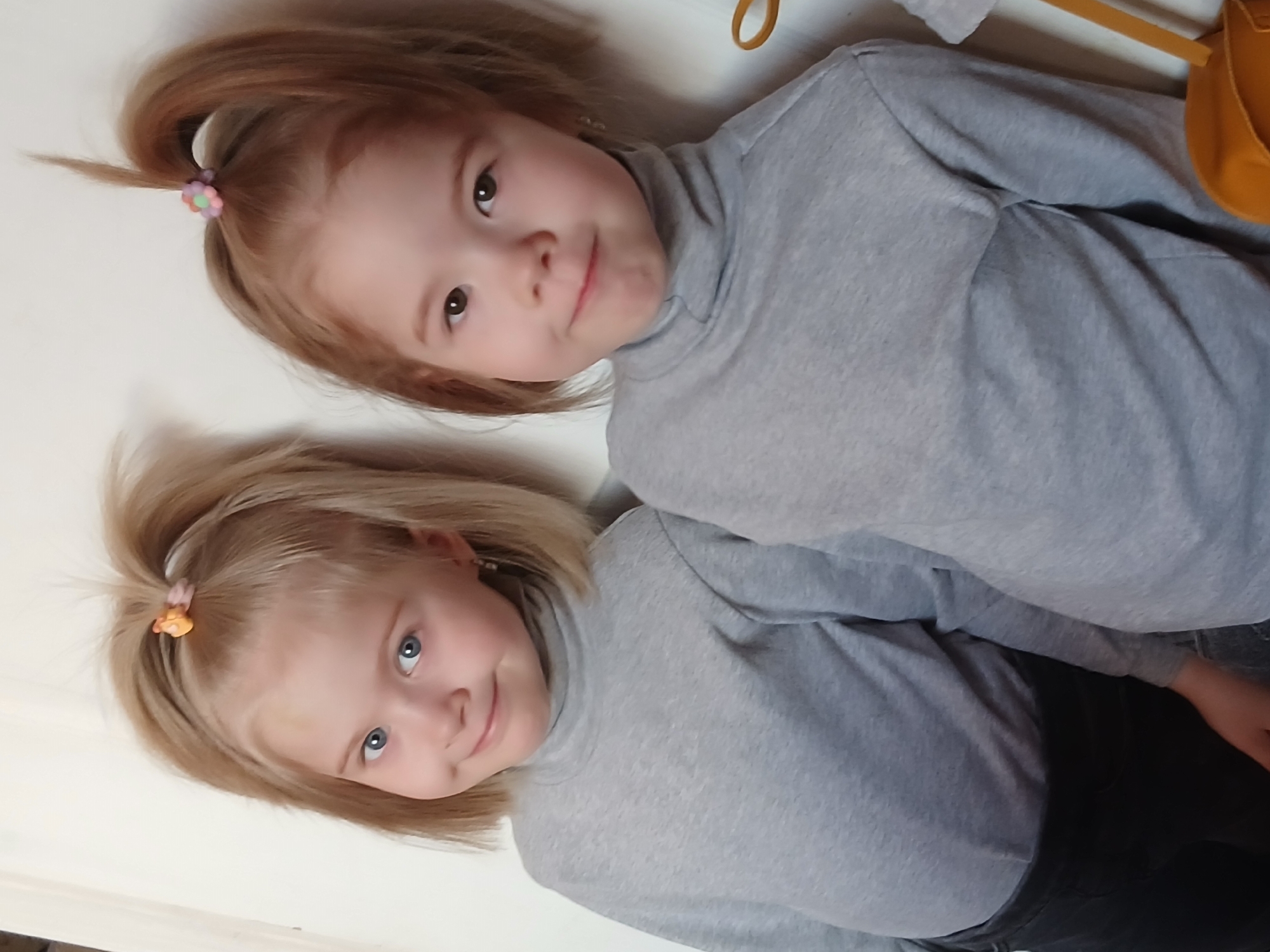 Two young girls, presumably sisters, are lying side by side on a white or light-colored surface. Both girls are wearing gray turtleneck shirts. The girl on the top has brown hair tied in a small ponytail with a flower-shaped hair tie. The girl on the bottom has blonde hair also tied in a small ponytail with an orange hair tie. They are both looking towards the camera and smiling faintly. The location is unknown, and there is no indication of Chaikovsky, Russia.