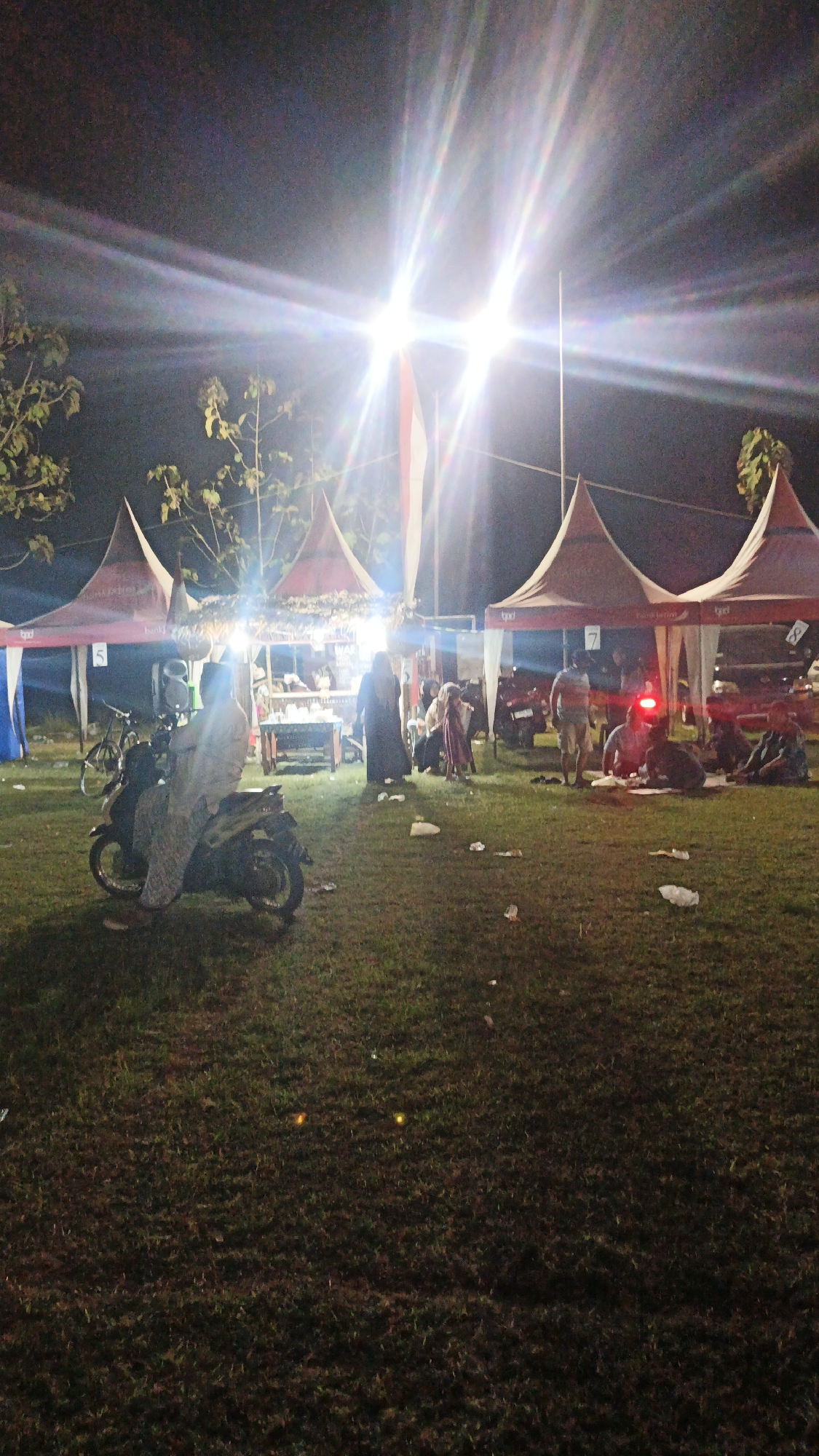 The image depicts an outdoor night market scene in Ketemasdungus, Indonesia, characterized by bright artificial lighting that creates lens flares. Several red and white tents are set up on a grassy field, serving as stalls. A person is seated on a scooter in the foreground, facing away from the camera. Behind the scooter, a stall is visible with people gathered around it, and more tents and people are spread throughout the background. The ground is covered in grass with some scattered litter. In the distance, what appear to be the headlights of a vehicle are illuminated.