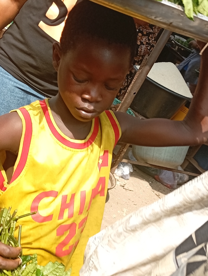 This image, set in Onitsha, Nigeria, depicts a young person positioned centrally in an outdoor market environment. The individual has dark skin and wears a yellow sleeveless top with red trim, featuring red lettering that includes "CHIA 27". Their head is tilted downwards, and their left hand holds a bundle of green leafy vegetables. Partially visible behind them is another individual wearing a black shirt with an orange design and blue denim. To the right, in the background, a black plastic basin contains a large mound of white grains, consistent with rice. Below this, the ground is unpaved dirt, with various items, some enclosed in clear plastic bags, scattered. A metallic bar extends horizontally across the top of the frame.