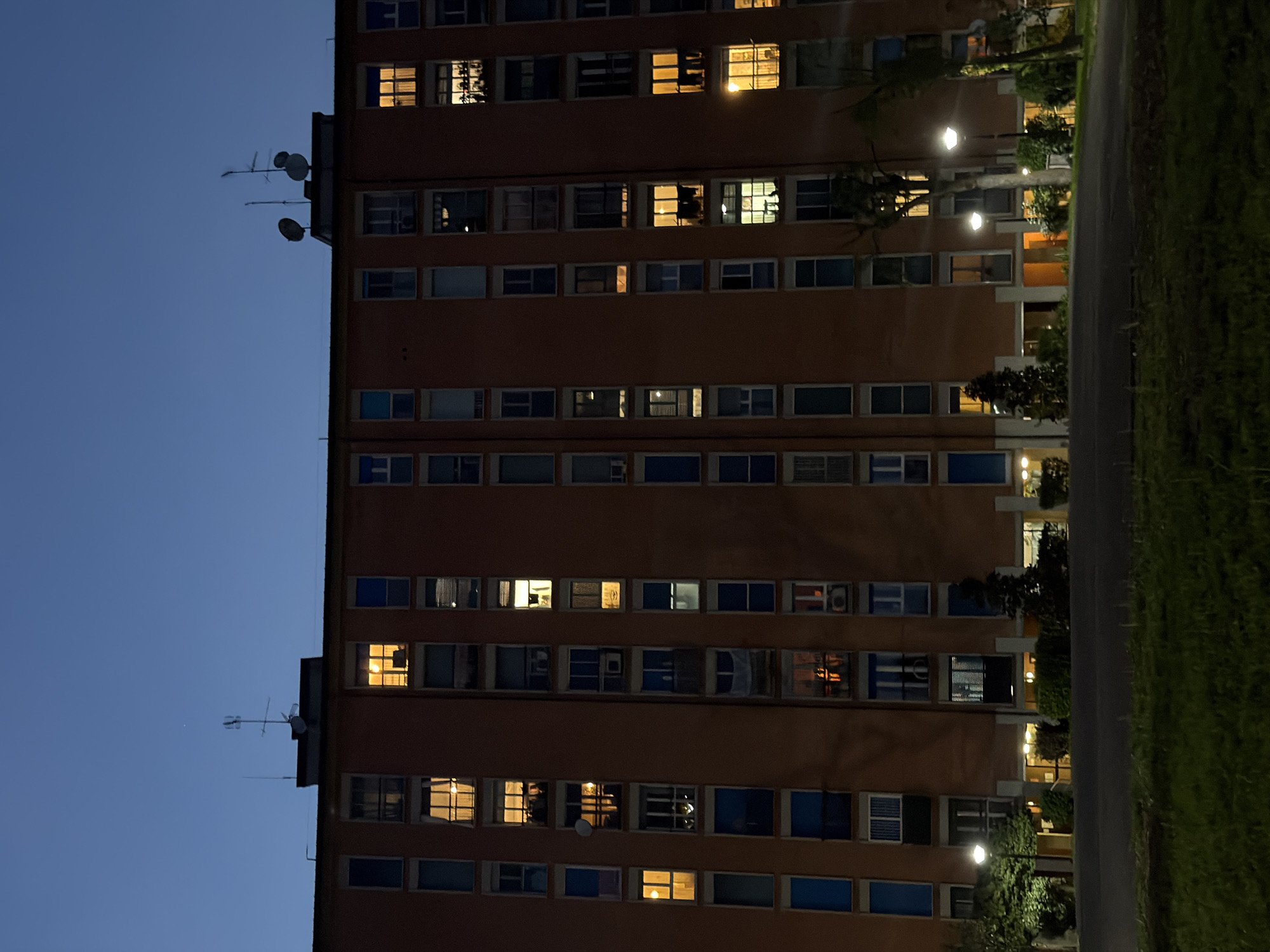 The image displays a tall, multi-story building at dusk or night in Milan, Italy. The building dominates the left and central parts of the frame, set against a clear, dark blue twilight sky. Its facade is a uniform reddish-brown or orange hue, featuring numerous rectangular windows arranged in orderly rows across multiple floors. Many windows emit a warm yellow or orange light from within, suggesting occupied interiors, while others remain dark. Some windows reveal glimpses of interior furnishings or blinds, and a few show air conditioning units.
On the building's flat rooftop, several antennas and satellite dishes are visible, silhouetted against the dark sky. To the right of the building, a paved pathway runs alongside a strip of green grass and scattered trees and bushes. Modern streetlights with hexagonal diffusers illuminate the ground level and the lower floors, casting pools of light.
The scene captures a peaceful urban evening. No individuals are visible, and no specific activity or event is taking place, beyond the implied domestic life within the illuminated apartments. The weather appears clear and calm. There is no visible text in the image.