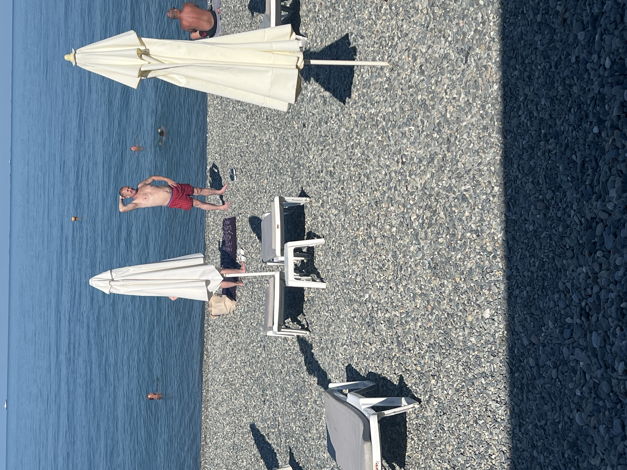 This image depicts a sunny day on a pebble beach in Sochi, Russia. In the background, the calm blue sea stretches to the horizon, with a distant white boat visible. Several people are in the water. In the foreground, two white beach umbrellas are prominent, casting shadows on the pebbled shore. Beach chairs are set up on the sand, with some occupied and some empty. One man in red checkered shorts stands on the beach, facing the water, with his hand behind his head. Another man, seen from behind, is seated on a beach chair. The overall scene conveys a relaxed summer atmosphere at a seaside resort.