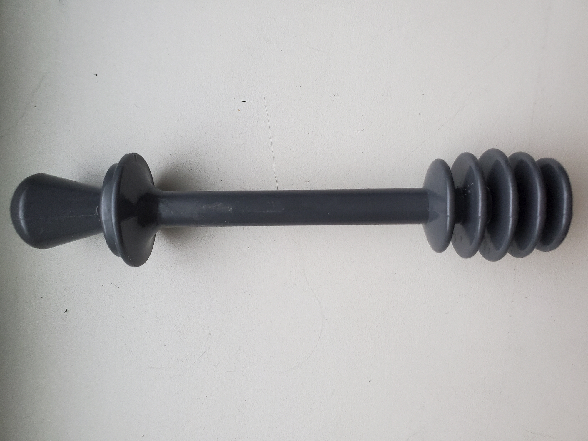 A dark gray plastic object, resembling a small plunger or stopper, is positioned vertically against a textured white surface. The object has a tapered handle at the top and a ribbed, disc-like base at the bottom. The background is a plain, off-white wall. There is no indication of the city of Tiraspol, Moldova, or any environmental context related to a specific location.