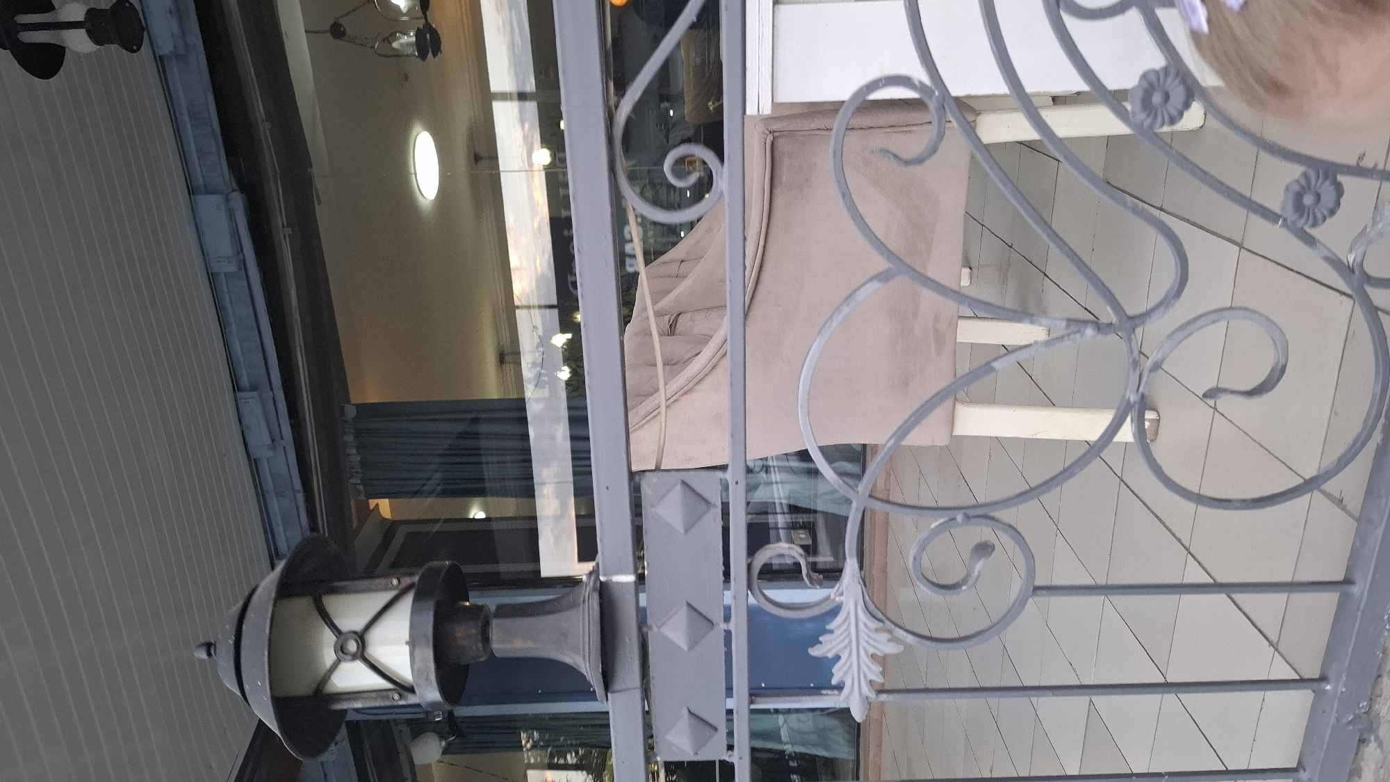 The image depicts an outdoor seating area with a lamppost and ornate metal railings in the foreground. Behind the railings, a light brown upholstered armchair is visible, positioned in front of a building with large glass windows. The building's interior, illuminated by ceiling lights and chandeliers, reflects the sky, which appears to be at sunset. The floor is tiled. While the specific location in Shymkent, Kazakhstan, is not identifiable from the visual elements alone, the scene suggests a cafe, restaurant, or a similar establishment in an urban setting.