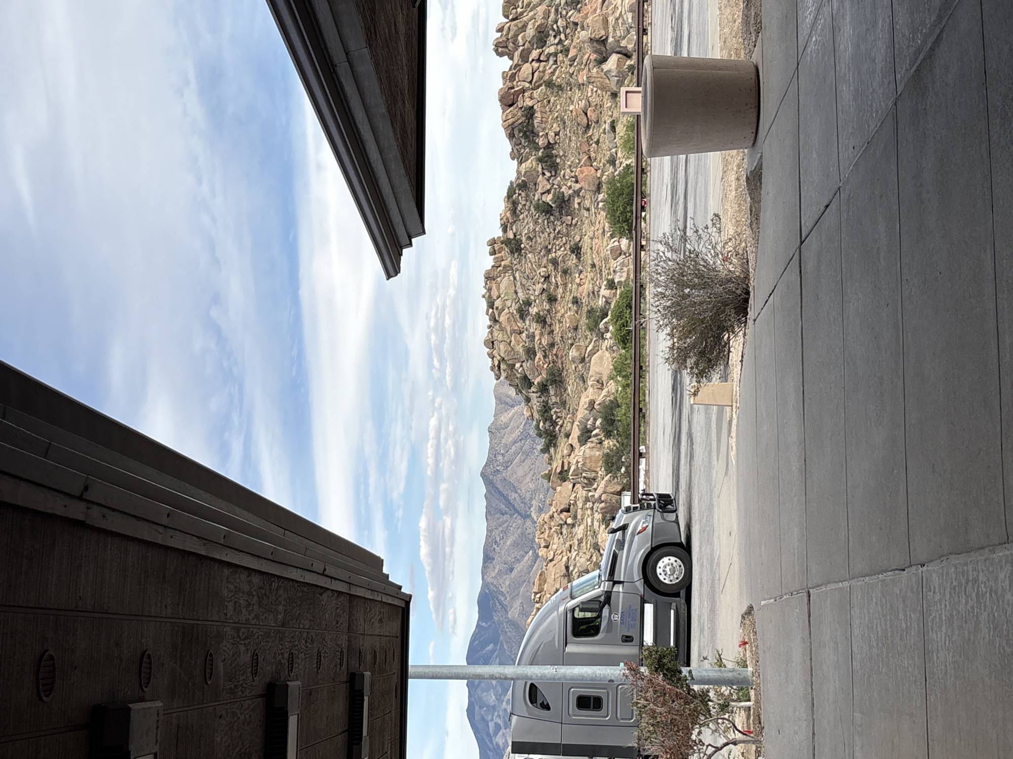 The image shows a gray semi-truck parked on the side of a road, with a mountainous landscape in the background. The truck is facing left, and its side is visible. The landscape consists of rocky hills with sparse vegetation. The foreground shows a concrete walkway and a large, cylindrical trash receptacle. The scene appears to be in an outdoor rest stop or parking area, likely in a desert or semi-arid region. There are two overhanging structures with wood-paneled ceilings and lighting fixtures, suggesting a covered area for vehicles or people. The sky is partly cloudy. The image is captured from a slightly elevated, ground-level perspective. The location is identified as Dragoon, United States.