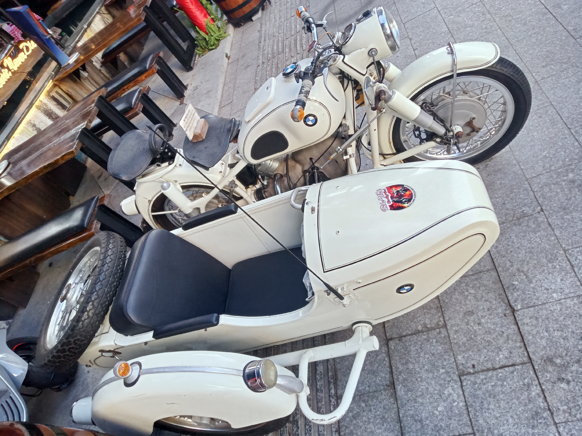 The image shows a vintage BMW motorcycle with a sidecar parked on a grey tiled surface. The motorcycle and sidecar are white. The sidecar has a black seat, a BMW emblem, and a decal featuring the text "BIKE VAT COLLECTION". The motorcycle is parked next to outdoor seating of a restaurant. The location appears to be a street in Nha Trang Ward, Vietnam.