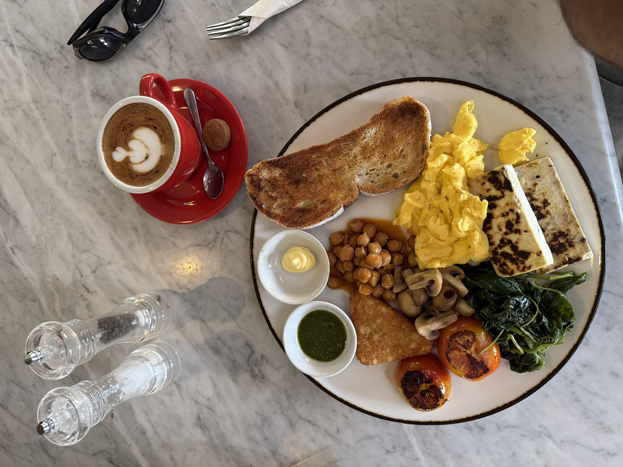 This media file presents an overhead view of a breakfast setting laid out on a white marble table, likely in a cafe or restaurant in North Kuta, Indonesia. The central focus is a large, oval-shaped white plate laden with a diverse selection of breakfast foods, accompanied by a coffee cup and other dining essentials.
On the main plate, there is a generous portion of scrambled eggs and a single, well-toasted slice of bread. Two rectangular pieces of grilled paneer or halloumi cheese are positioned next to the eggs. Other items include baked chickpeas (resembling baked beans), a serving of sautéed green leafy vegetables (possibly spinach), grilled mushrooms, and two halves of grilled tomatoes. A hash brown is also visible. Two small white condiment bowls sit on the plate: one contains a dollop of a pale yellow creamy sauce (possibly mayonnaise), and the other holds a vibrant green chutney-like sauce.
To the left of the breakfast plate is a bright red coffee cup and saucer. The cup contains a latte with delicate white latte art, resembling a heart or a stylized swan, on its dark brown surface. A small, round, brown biscuit rests on the saucer next to a spoon.
Further details on the table include a pair of black sunglasses, a silver fork, and two clear glass salt and pepper grinders. The grinders show visible coarse salt crystals and black peppercorns within. The bright, even lighting suggests it is daytime. The overall scene depicts a prepared meal, implying an activity of dining or about to dine. No specific text is visible within the image.