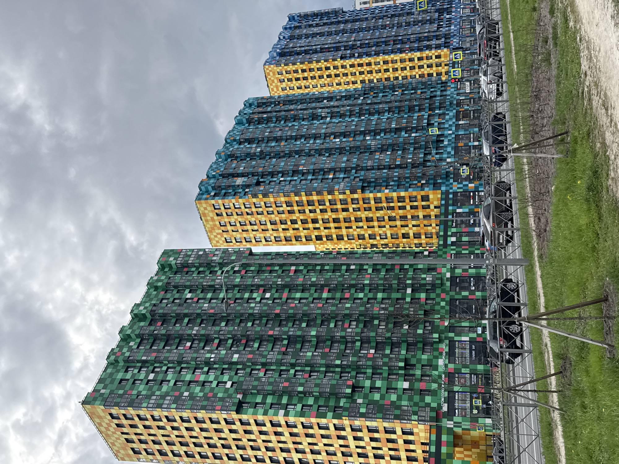 Three tall apartment buildings with pixelated facades in green/yellow, teal/yellow, and blue/yellow stand next to each other. Cars are parked along the side of the road next to the buildings, separated by a low fence. The sky is cloudy. The location is likely Saint Petersburg, Russia, based on the architecture and style of the buildings.