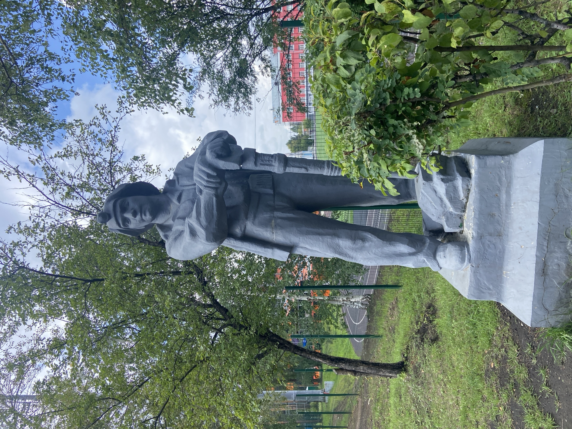 A gray, concrete statue of a miner stands on a pedestal in an outdoor, grassy area with trees. The miner wears a helmet and work clothes, and holds a mining tool. In the background, a red brick building with white windows is visible, along with a green fence and a running track. The statue is located in the city of Cheremkhovo, Russia, specifically at the Cheremkhovo Mining and Construction College.