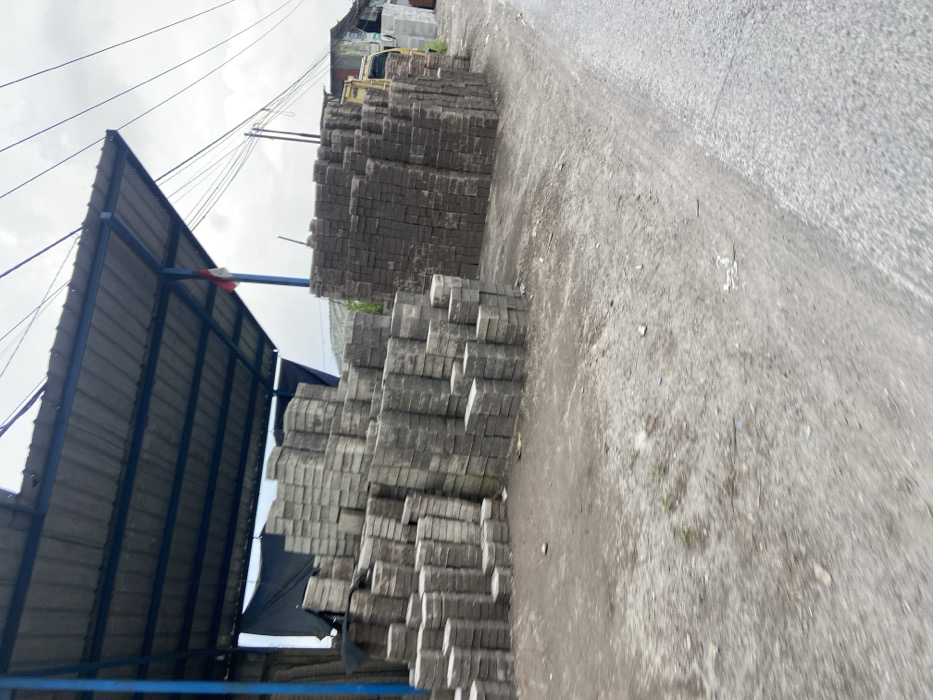 The image shows a pile of concrete bricks stacked on a dirt road in Kecamatan Margaasih, Indonesia. The bricks are arranged in a neat, organized manner, with some bricks stacked higher than others. There is a blue metal awning above the bricks, and a white truck is partially visible in the background. The road is made of asphalt and is relatively narrow, with a dirt shoulder on one side. There are no people in the image.