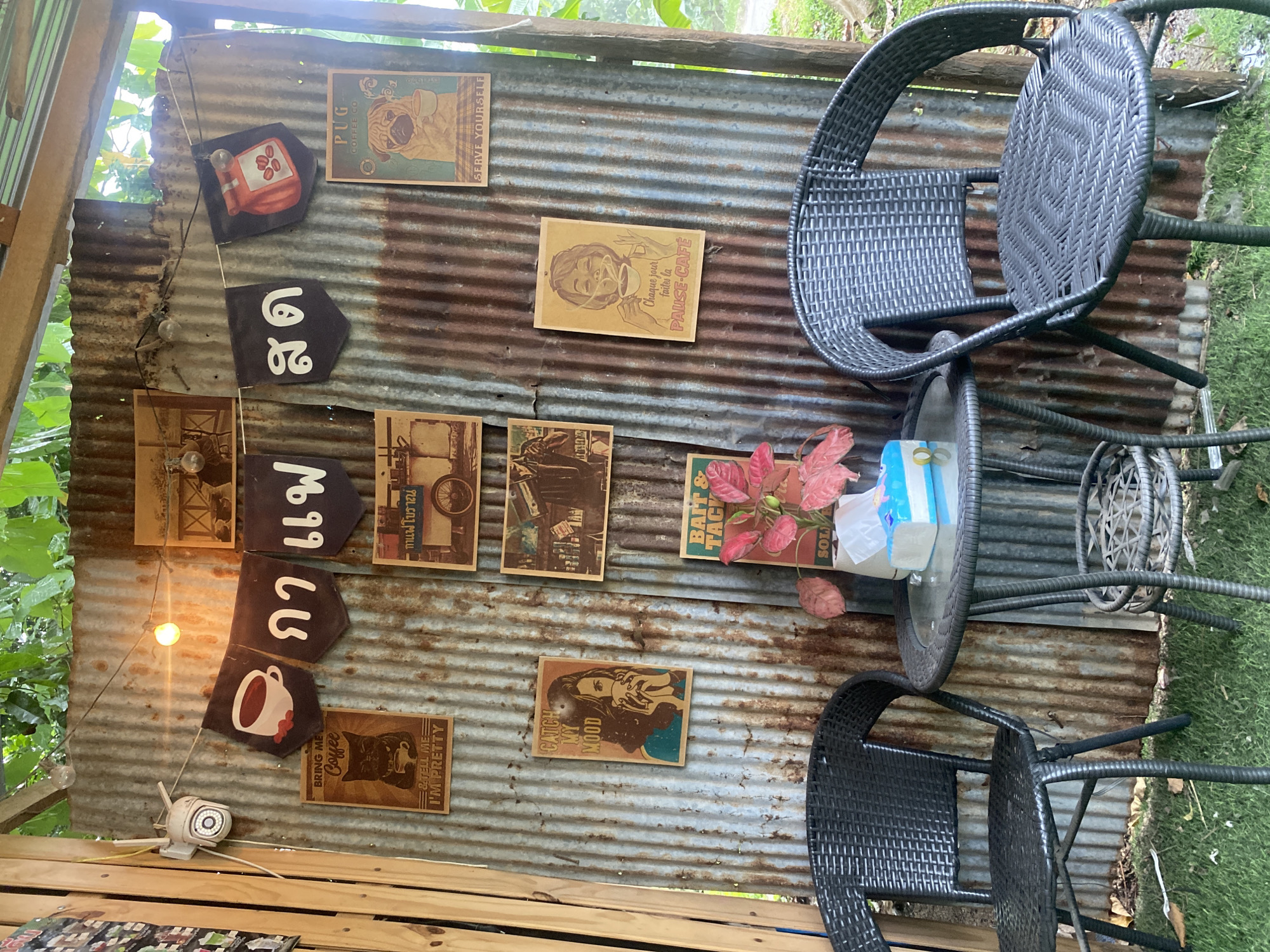 The image depicts an outdoor seating area with two black wicker chairs and a small round glass-topped table set on a patch of green grass in front of a corrugated metal wall. The wall is adorned with several vintage-style posters and banners. One banner features a drawing of a coffee cup with red beans, and other banners display Thai script. The posters include images of a black cat with a coffee cup, a woman drinking coffee, a pug, and a woman with a steaming cup of coffee captioned in French. A tissue box sits on the table, along with a small potted plant with red leaves. A string of bare lightbulbs is strung across the top of the metal wall, with one bulb illuminated. A security camera is mounted on a wooden post to the left of the wall. The environment appears to be a casual outdoor cafe or restaurant. The mention of Baan Lamai, Thailand suggests this is likely the location of the establishment.