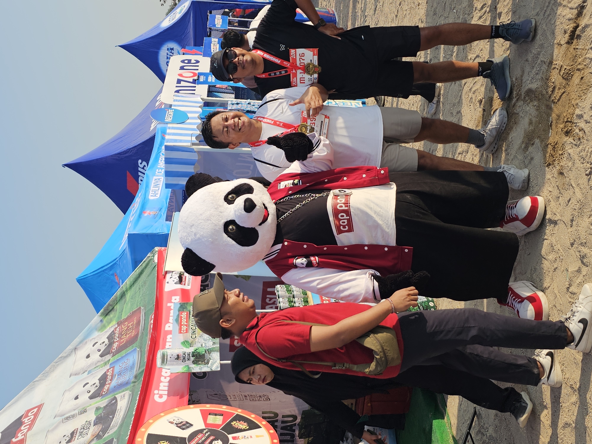 At an outdoor event, likely a race finish line, in Denpasar, Indonesia, a group of people pose for a photograph under a clear sky with tents in the background. A person in a panda mascot costume stands in the center, wearing a red and white jacket with "cap panda" written on it, and a silver chain. To the right of the mascot, two men proudly display finisher medals. One man, wearing a white t-shirt and light-colored shorts, gives a thumbs-up and smiles. The other, in a black t-shirt and black shorts, also smiles and holds his medal. To the left of the mascot, a person wearing a red polo shirt and a green crossbody bag stands with their arms at their sides, while a person in a black hijab stands behind them. In the background, several tents are visible, some with branding for "cap panda" products, and blue tents with logos, suggesting corporate sponsorships. The ground is sandy, with some water stains visible. The overall atmosphere appears celebratory, with participants and attendees enjoying the post-race activities.