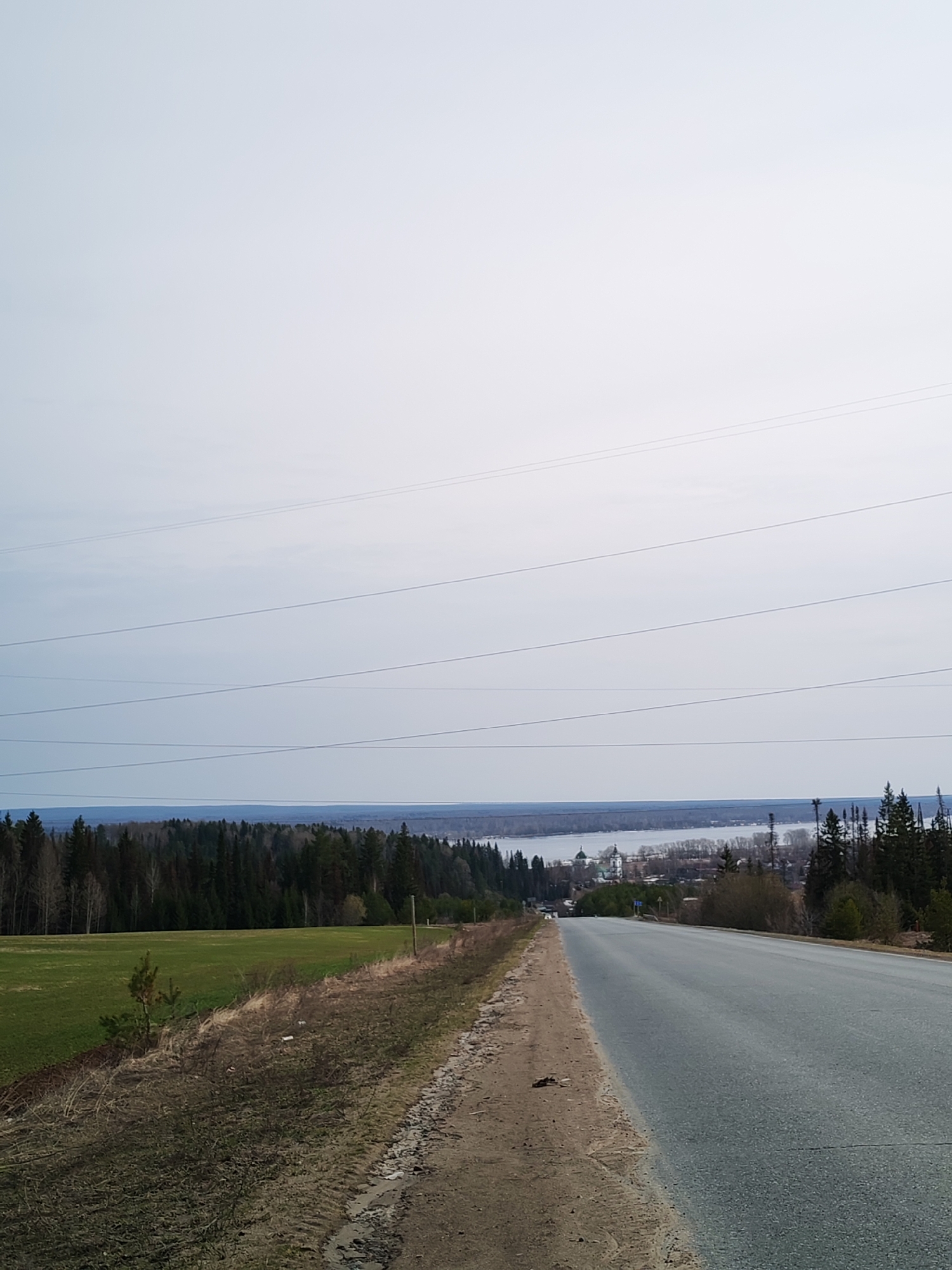 The image depicts a gray asphalt road leading downhill towards a distant settlement in Нечкино, Russia. The sky is overcast. Power lines stretch across the top portion of the image. To the left of the road is a field of green grass transitioning into barren earth, while to the right is a dirt shoulder with scattered vegetation and trees. In the distance, a body of water is visible beyond a dense forest, adjacent to a village featuring what appears to be a church or religious structure with a green dome.