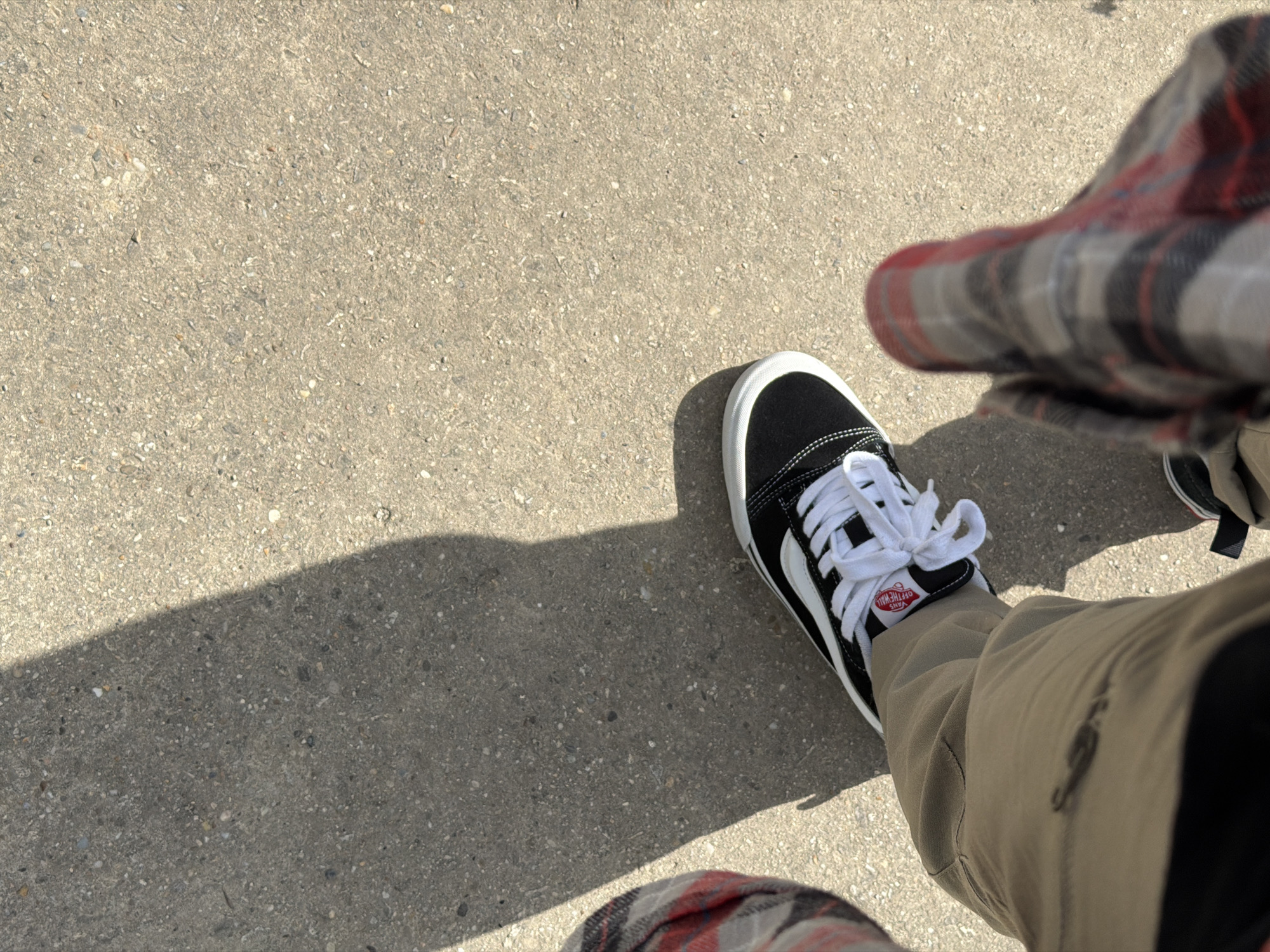 The image shows a person's legs and feet from a first-person perspective, standing on a concrete surface. The person is wearing tan pants and black Vans sneakers with white laces and a red Vans logo tag. A shadow of the person is cast on the concrete. The person is also wearing a red, grey, and black plaid shirt or jacket. The environment appears to be outdoors, possibly a sidewalk or path. There is no identifiable information in the image that would suggest a specific location in Vienna, Austria.