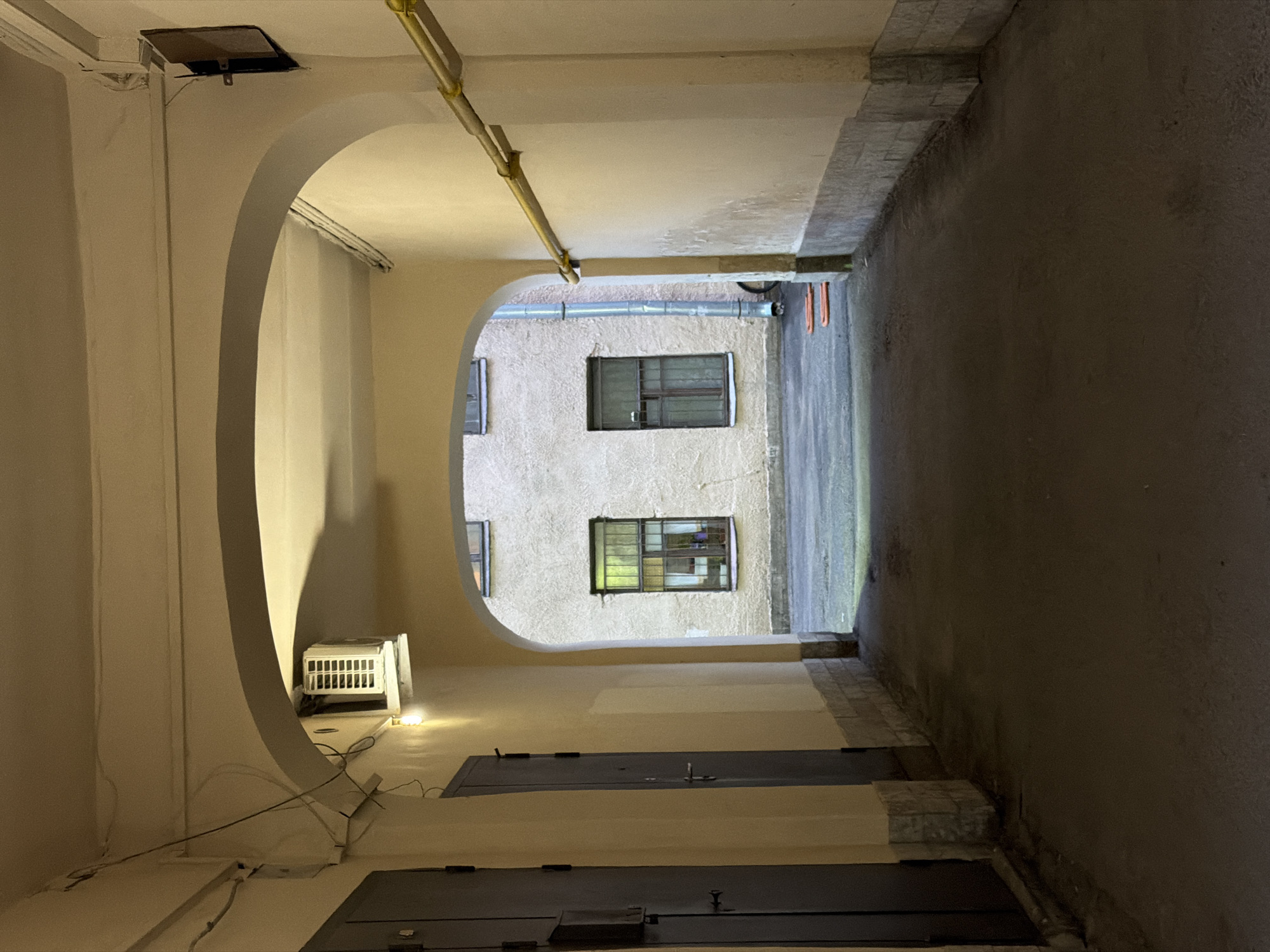 The image depicts an arched passageway within a Saint Petersburg courtyard. The passageway walls are painted a pale yellow, with the lower section adorned with a stone-like pattern. Two dark grey doors are visible on the left side of the passageway. An air conditioning unit is mounted high on the left wall. The passageway opens into a wider courtyard area where the facade of a building is visible, featuring two windows with barred panes. A downspout runs vertically beside the windows, and a yellow pipe is mounted horizontally along the right wall of the passageway. The ground is paved with asphalt. The scene is dimly lit, suggesting it is either evening or night.