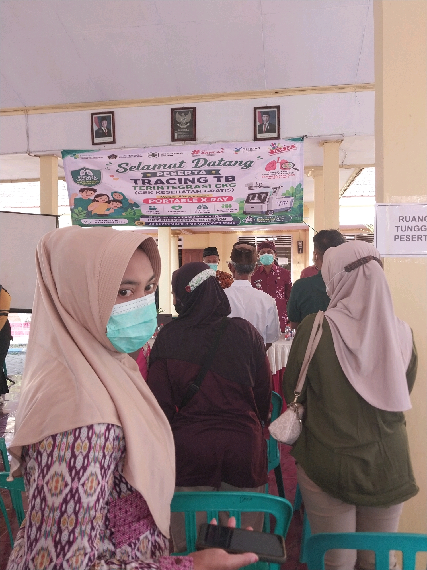 A woman wearing a light brown hijab and a blue surgical mask is in the foreground, looking to her left. She is wearing a patterned dress with purple, pink, and white geometric designs. Behind her, other people are also wearing masks and hijabs, including a woman in a dark maroon hijab and a woman in a light olive green jacket. In the background, a banner promotes a tuberculosis (TB) tracing event. The banner is in Indonesian and features text such as "Selamat Datang" (Welcome) and information about tracing TB. There are framed portraits on the wall above the banner. The setting appears to be an outdoor or semi-outdoor covered area, possibly a community hall or gathering space, with light-colored walls and pillars. The lighting suggests it is daytime. The overall scene depicts a health outreach event.
