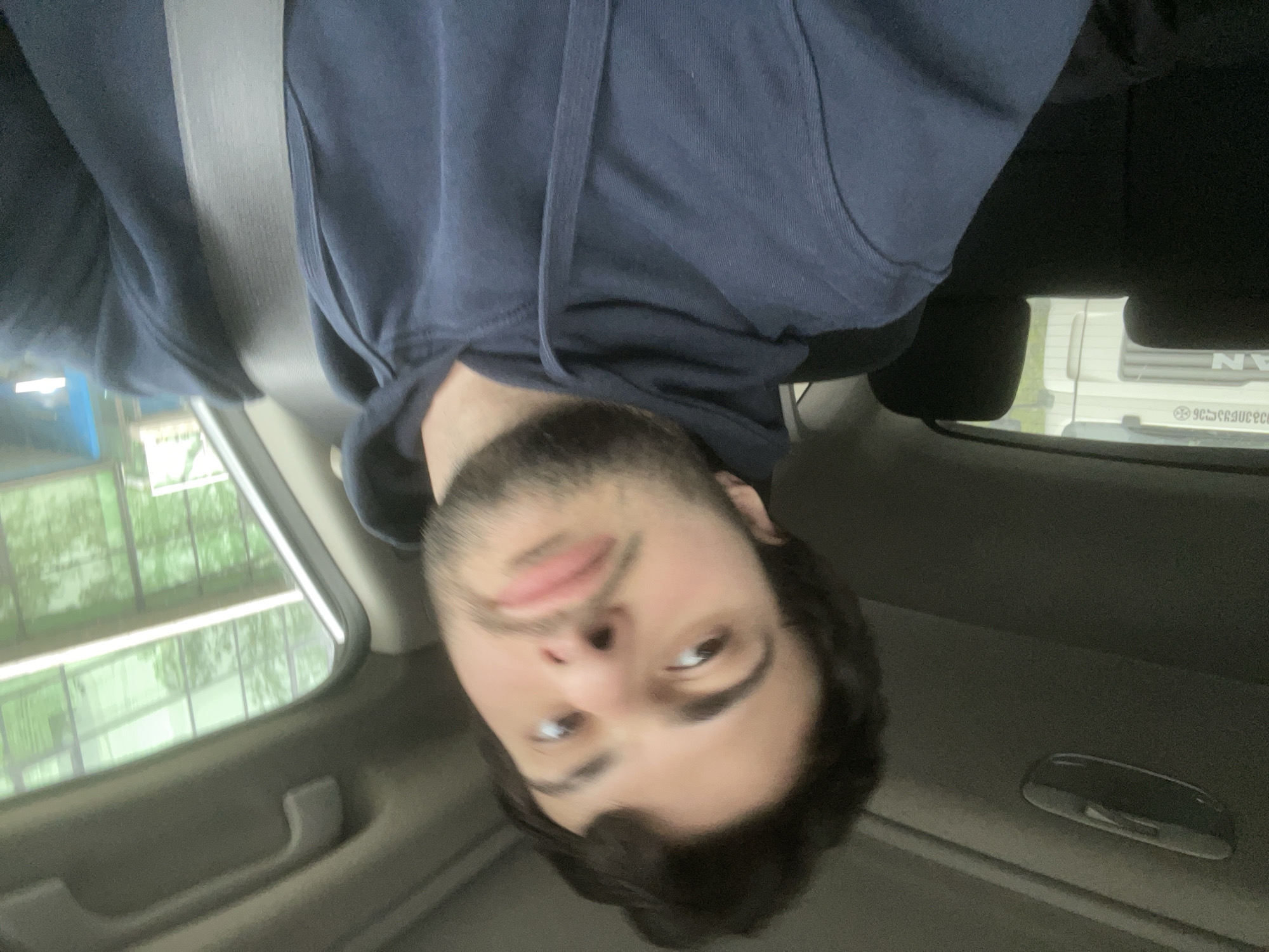A person wearing a navy blue hooded sweatshirt is inside a vehicle. The person is wearing a seatbelt. A building with several windows is visible through the vehicle's window, and the word "VAN" is visible on another vehicle outside. Based on the reversed text on the other vehicle's window and the shape of the letters, the vehicle is likely displaying Georgian text. This suggests that the location is Tbilisi, Georgia.