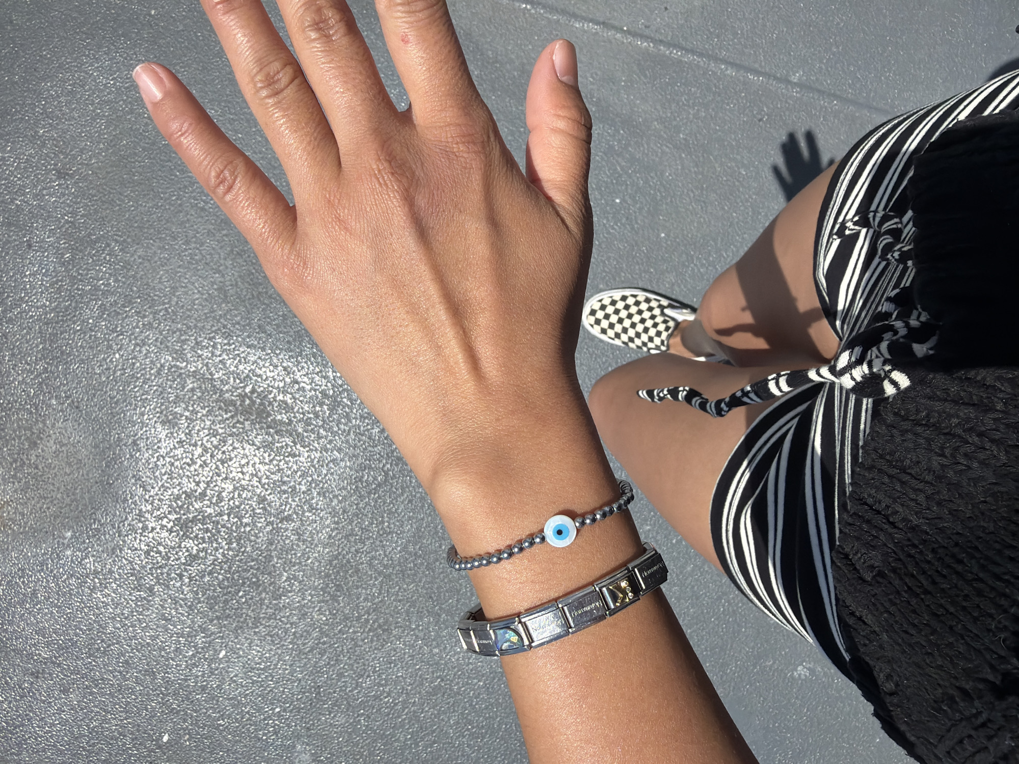 In Point Roberts, United States, a close-up shot captures a person's arm and hand from a first-person perspective. The individual is wearing two bracelets on their left wrist. The first bracelet, closer to the hand, is a silver chain with small, dark beads and a central charm resembling an evil eye with a white iris and blue pupil. Adjacent to this, on the forearm, is a silver link bracelet adorned with various charms, including one with a colorful image and others with text or geometric designs. The person's hand is open, fingers extended, and positioned in the foreground. Their lower body is also visible, showing a black top and a striped skirt with alternating black and white lines. A black and white checkered slip-on shoe is visible on their right foot. The ground beneath them appears to be a textured, gray surface.