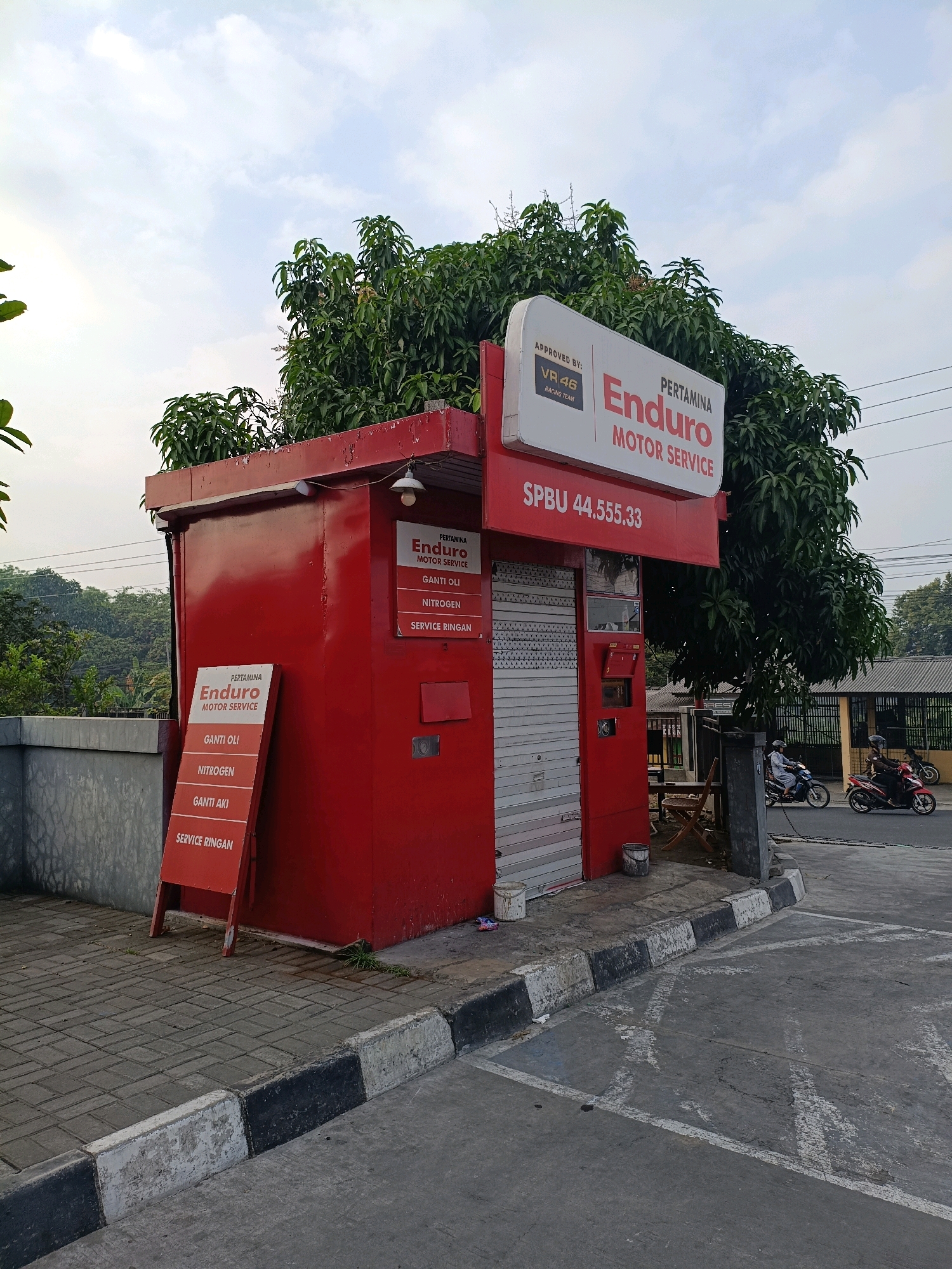 The image shows a red Pertamina Enduro Motor Service booth in Maguwoharjo, Indonesia. The booth has a white sign with red lettering that reads "PERTAMINA ENDURO MOTOR SERVICE" and "APPROVED BY: VR46 RACING TEAM". There is also a smaller sign to the left of the booth that lists services such as "GANTI OLI", "NITROGEN", "GANTI AKI", and "SERVICE RINGAN". In the background, there are trees and power lines, and two people on motorcycles are riding on the street. The ground is paved with cobblestones and concrete.