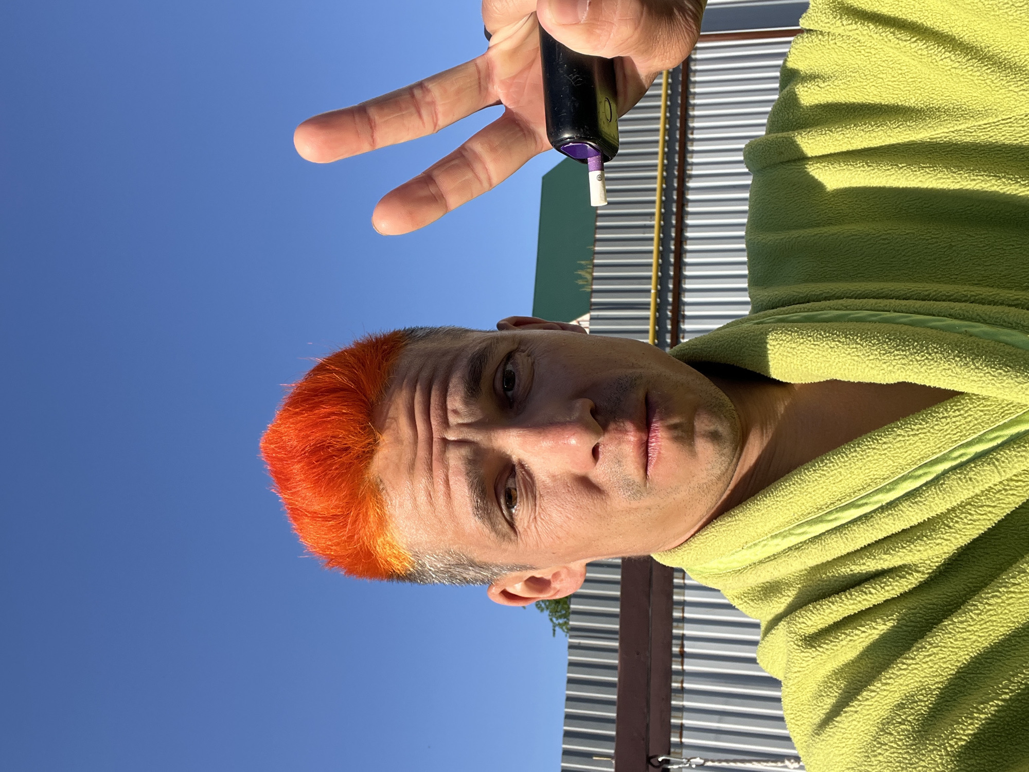 The image is an outdoor shot of a man with orange hair, dressed in a light green, zippered fleece jacket or bathrobe, holding a black device in his hand. His hand is positioned to display three fingers. The backdrop includes a clear blue sky and a section of a corrugated metal structure, likely a fence or wall. Based on available information, the location is Chaikovsky, Russia. The lighting suggests it is daytime. The man is facing the camera with a slightly furrowed brow.